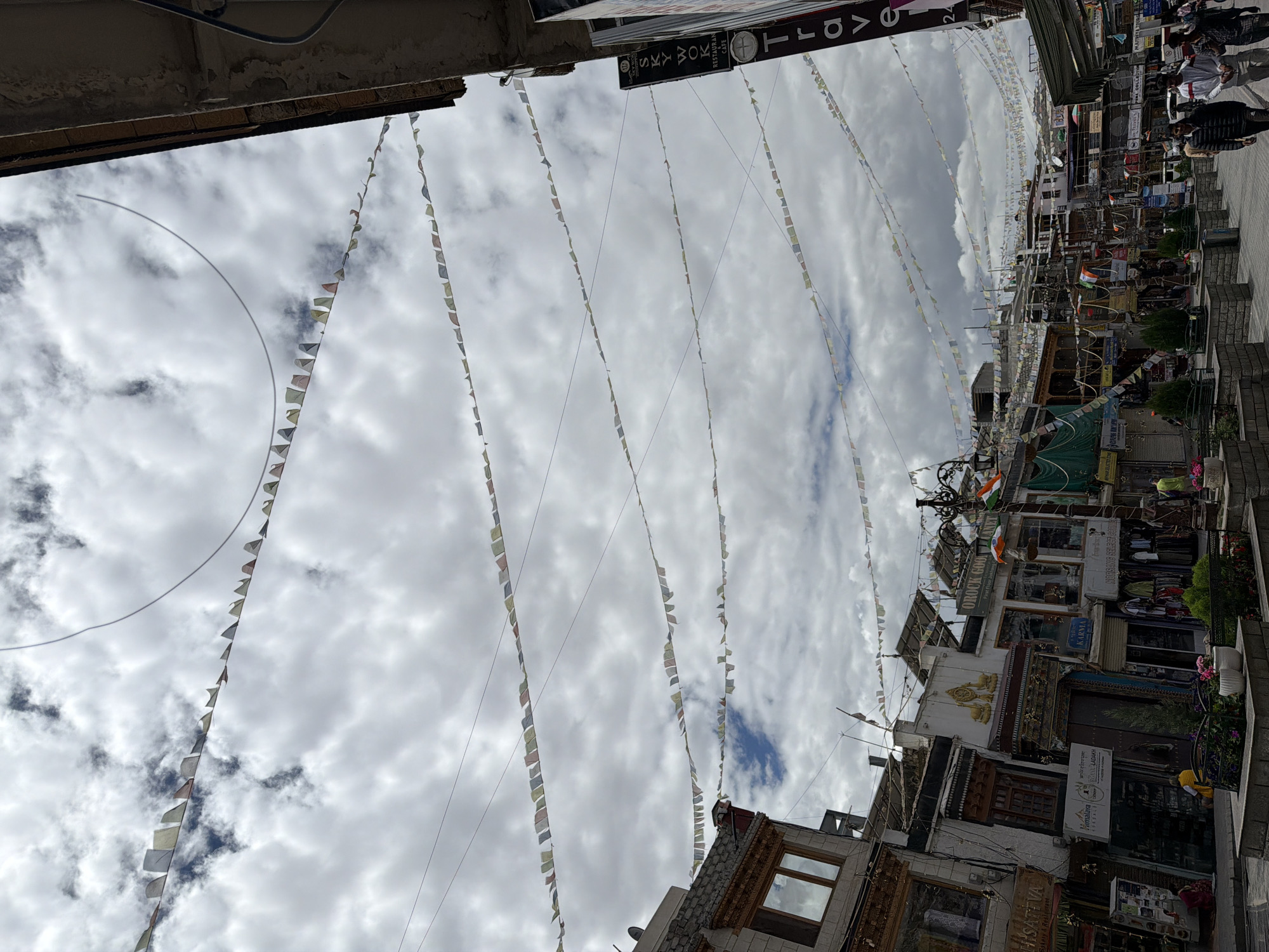 The image shows a street scene in the city of Leh, India. Prayer flags are strung across the sky, which is overcast with clouds. Buildings line the street, with shops and businesses visible. Several people are walking on the sidewalk. The architecture appears to be traditional Ladakhi style with influences from Tibetan Buddhism. The overall impression is of a bustling market street in a mountainous region. There is a sign that says "SKY WOK RESTAURANT CAFE" and another that says "TRAVEL".