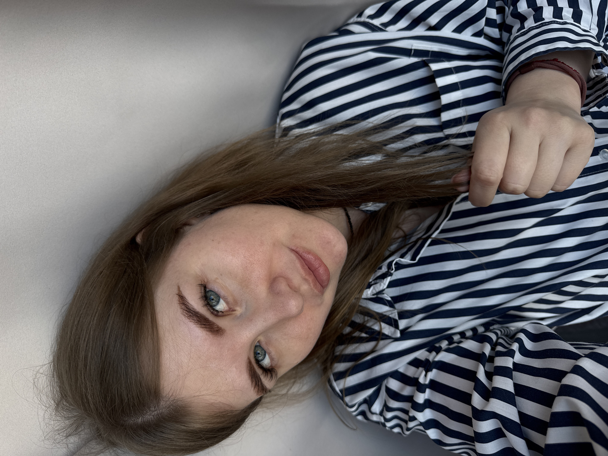A young woman lies on a light-colored surface, likely a couch or bed, wearing a blue and white striped shirt. She is looking directly at the camera with a slight smile. Her hair is light brown and her eyes are blue. Her right hand is visible holding a strand of her hair. No specific details indicate the city of Gornyi or its surroundings.