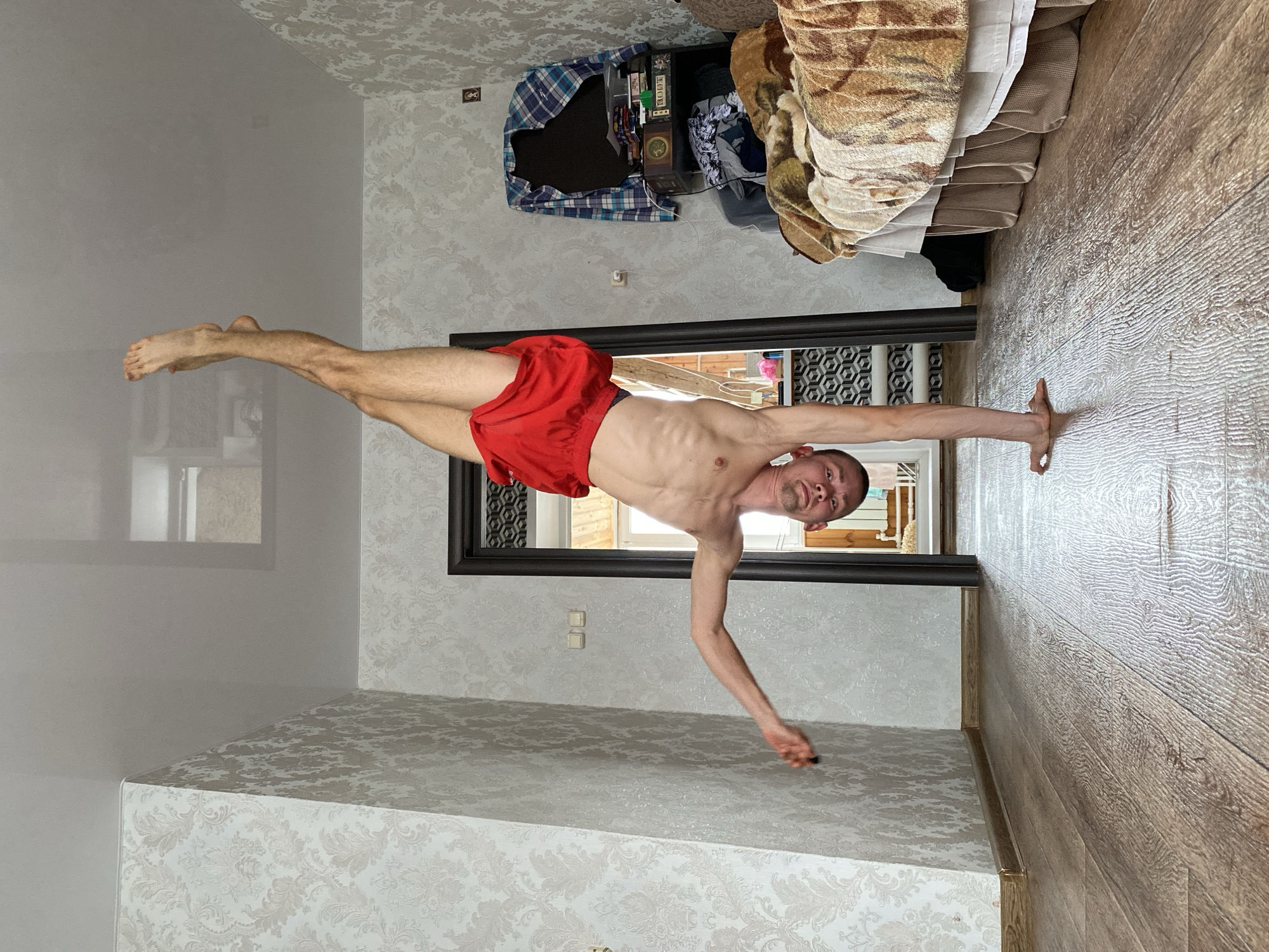 A shirtless man in red shorts is performing a one-arm handstand in what appears to be a domestic room. His left hand is flat on a light wood laminate floor, supporting his entire body, while his right arm hangs loosely, bent at the elbow. His left leg is extended straight upwards, and his right leg is bent at the knee. He has a muscular physique and a focused expression.
The room features light-colored walls adorned with a subtle, patterned wallpaper. A dark-framed doorway is visible behind the man, leading into another brightly lit room that appears to have lighter walls and a window. In the background, to the left of the doorway (when oriented correctly), there is a cluttered area with clothes, including a blue and white plaid shirt, and various items on what looks like a shelf or top of a piece of furniture. A brown, patterned blanket or textile is also present in this area. Electrical outlets are visible on the wall. The room is brightly lit, suggesting it is daytime. On some items in the cluttered background, partial Cyrillic text "МООТ" and "СМЕ" can be seen. The location context for this scene is Ulyanovsk, Russia.