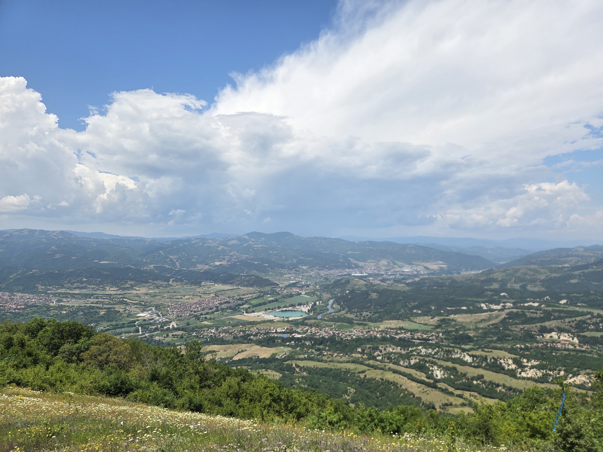This panoramic image depicts a vast landscape viewed from an elevated position, looking down over the city of Brejani, Bulgaria. The upper half of the scene is dominated by a bright blue sky partially covered with large, fluffy cumulus clouds. The clouds are predominantly white, with denser formations on the left and more scattered, slightly grayer clouds extending towards the right horizon.
In the immediate foreground, the sloping hillside is covered with vibrant green grass and a diverse array of wildflowers, primarily white and yellow, with some hints of purple. Below this floral carpet, a dense band of dark green deciduous trees and bushes extends into the midground.
The midground reveals a sprawling valley containing the settlement of Brejani. Numerous buildings with reddish-brown tiled roofs are visible, interspersed with agricultural fields ranging from lush green to lighter brown and yellow. A prominent, irregularly shaped light blue body of water, possibly a lake or reservoir, is centrally located within the valley. A winding river, appearing dark green or blue, flows through the valley, passing near the settlements and fields. Various roads and pathways are discernible, traversing the landscape and connecting different parts of the urban and rural areas. On the right side of the midground, distinct terraced hillsides with exposed light-colored earth (possibly eroded clay or sand formations) contrast sharply with the surrounding green vegetation. A thin blue ribbon or strip of fabric is visible amongst the foliage on the far right.
In the background, successive layers of hills and mountains extend towards the distant horizon, gradually fading into a hazy blue due to atmospheric perspective, culminating in a faint mountain range beneath the distant clouds. There are no visible people or discernible actions in the image.