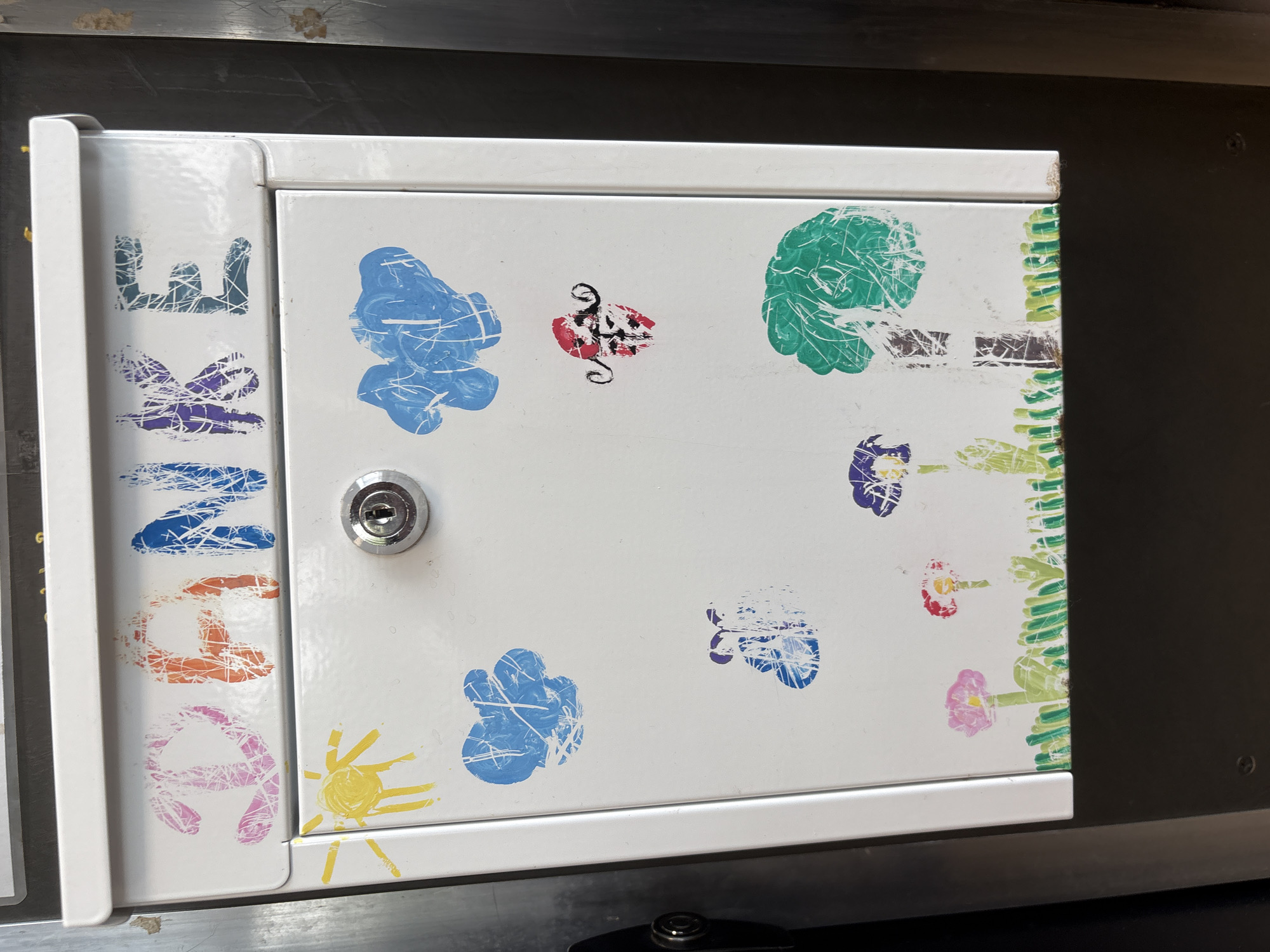 A close-up shot depicts a white metal cabinet or box with playful, amateurish decorations. The door is adorned with various hand-printed shapes and designs in different colors. These include blue cloud-like shapes, a green circle, a yellow sun, purple and red shapes that could be flowers or abstract designs, and a vertical strip of green marks on the right edge. The word "MENU" is printed vertically down the left side, with each letter in a different color. A metal keyhole is centered on the door. The box is located within a restaurant named "Ristorante Da Pino" in Kaarst, Germany.