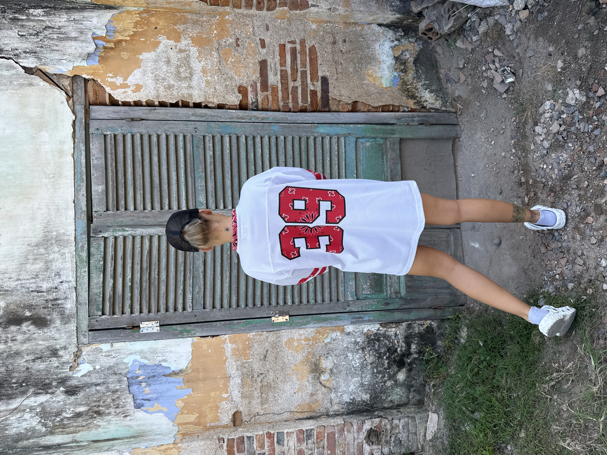 A person wearing a white jersey with the number 39 on the back, a black baseball cap, and white sneakers stands facing away from the camera in front of a weathered, green wooden door. The person has blonde hair pulled back and a tattoo on their left calf. The background consists of a crumbling, textured wall with visible bricks and peeling paint in shades of yellow, orange, and blue. The ground is covered with gravel, dirt, and overgrown grass. Debris, including broken bricks, is scattered to the right of the door.