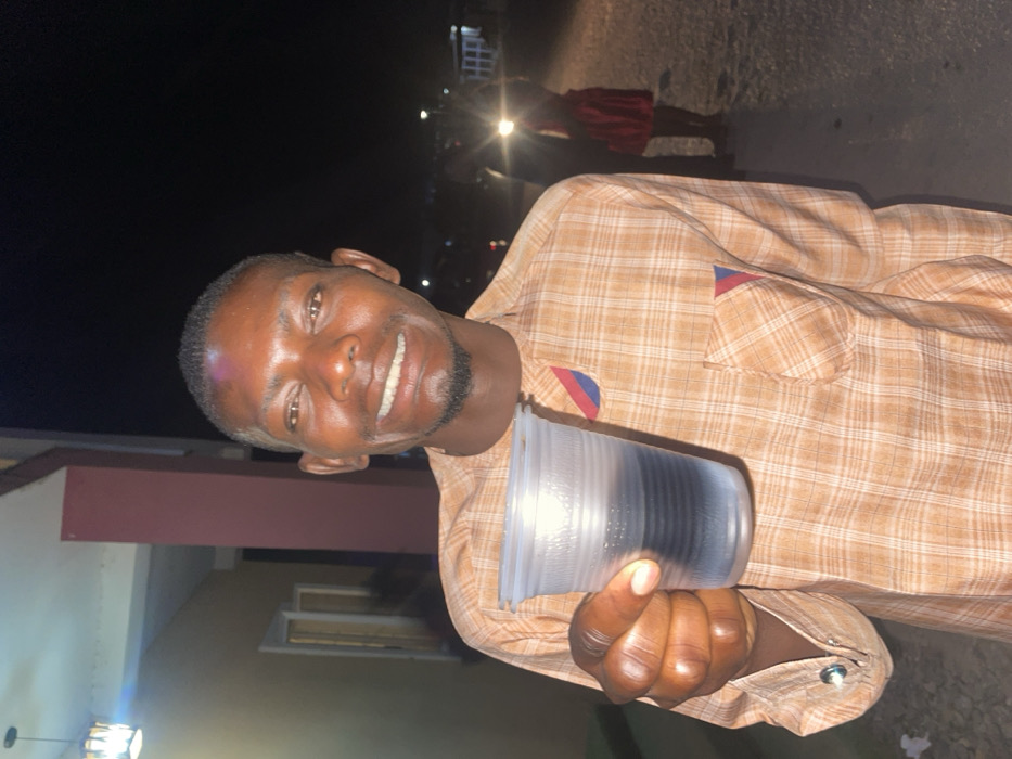 A smiling dark-skinned man holds a stack of clear plastic cups. He is wearing a light orange and white plaid shirt with a pocket. Red and blue striped fabric is attached to the shirt at the pocket and wrist. The background appears to be an outdoor nighttime setting. There is a red column behind the man, and a building with windows. A blurry figure in a red dress stands in the distance, with streetlights and dark sky visible overhead.