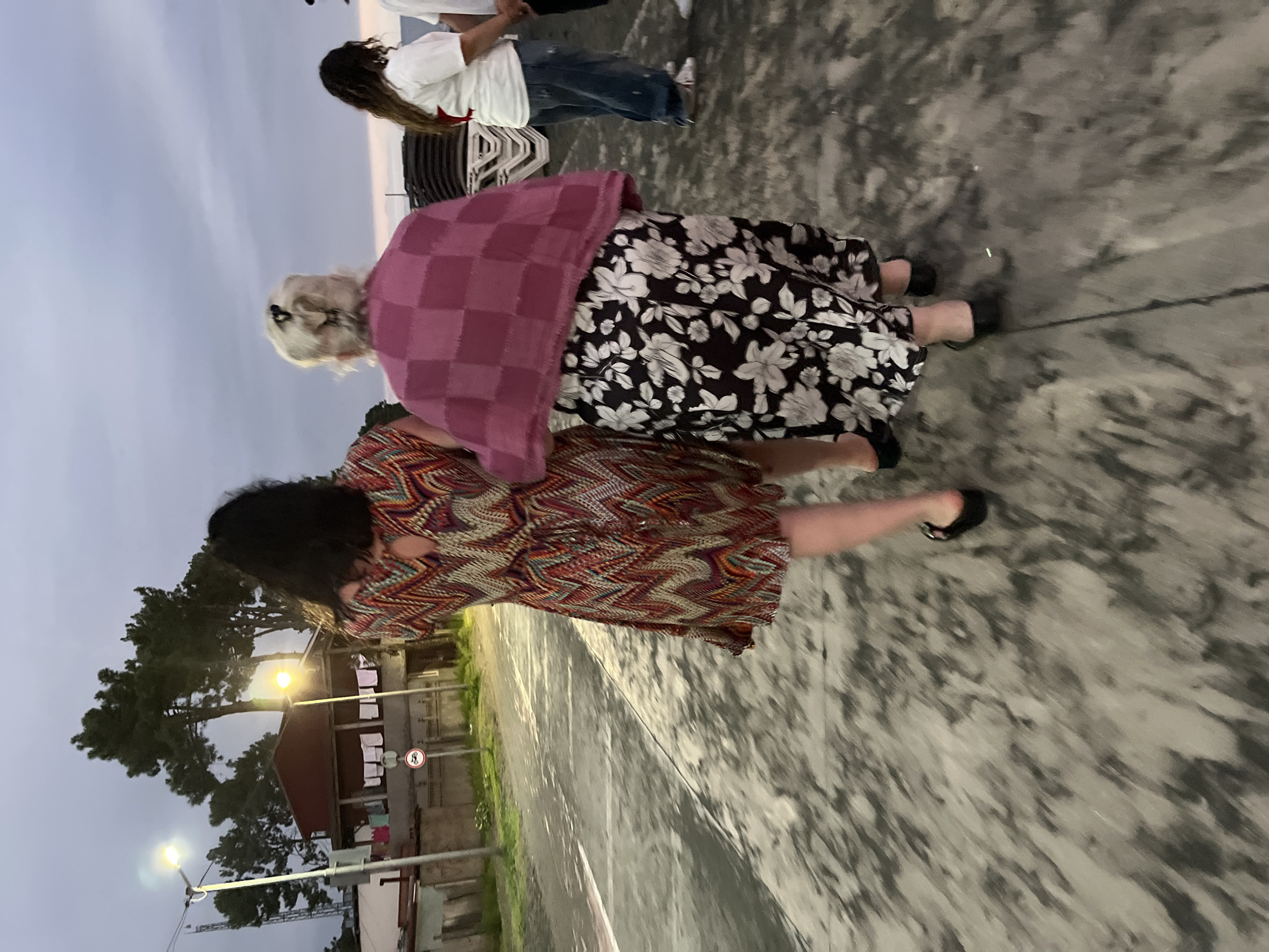 The image captures two women walking away from the camera on a paved surface, likely a sidewalk or promenade, in Ureki, Georgia. The sky above is overcast, with a mix of grey and pale pink hues suggesting either early morning or late evening. In the background, there is a building with a reddish-brown roof and laundry hanging outside. Several trees, including what appear to be tall pine trees, are visible. Streetlights illuminate the scene.
The woman in the foreground on the left is wearing a short, brightly colored dress with a zig-zag pattern in red, orange, yellow, blue, and white. She has long, dark hair. The woman next to her on the right is wearing a pink checkered shawl or wrap over a long black skirt with a white floral print. Her hair is styled in a blonde bun. Both women are wearing dark-colored flat shoes.
Further in the background, to the right of the two women, another person wearing a white t-shirt and blue jeans is visible, along with what appear to be stacked beach chairs. The overall impression is a casual, outdoor setting in a coastal town.