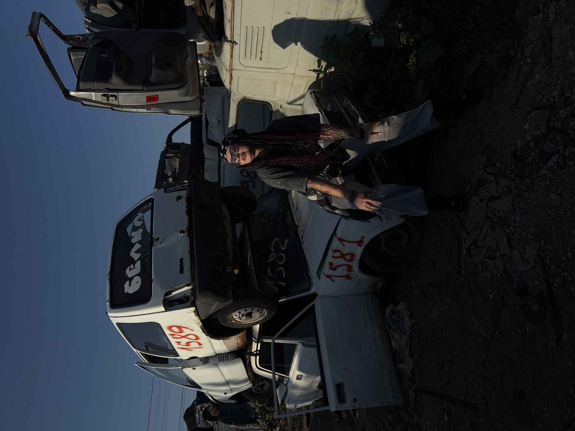 The image depicts a woman sitting on a wrecked car in a junkyard, likely in Tolyatti, Russia, known for its automotive industry and associated scrap yards. The woman, dressed in a casual outfit, is positioned on the hood of a white car that is stacked on top of another white car. Both vehicles appear heavily damaged and abandoned. The background consists of a clear blue sky, other wrecked vehicles, and some overgrown vegetation. The lighting suggests it's either early morning or late afternoon, casting long shadows. The overall scene conveys a sense of urban decay and post-industrial landscape.