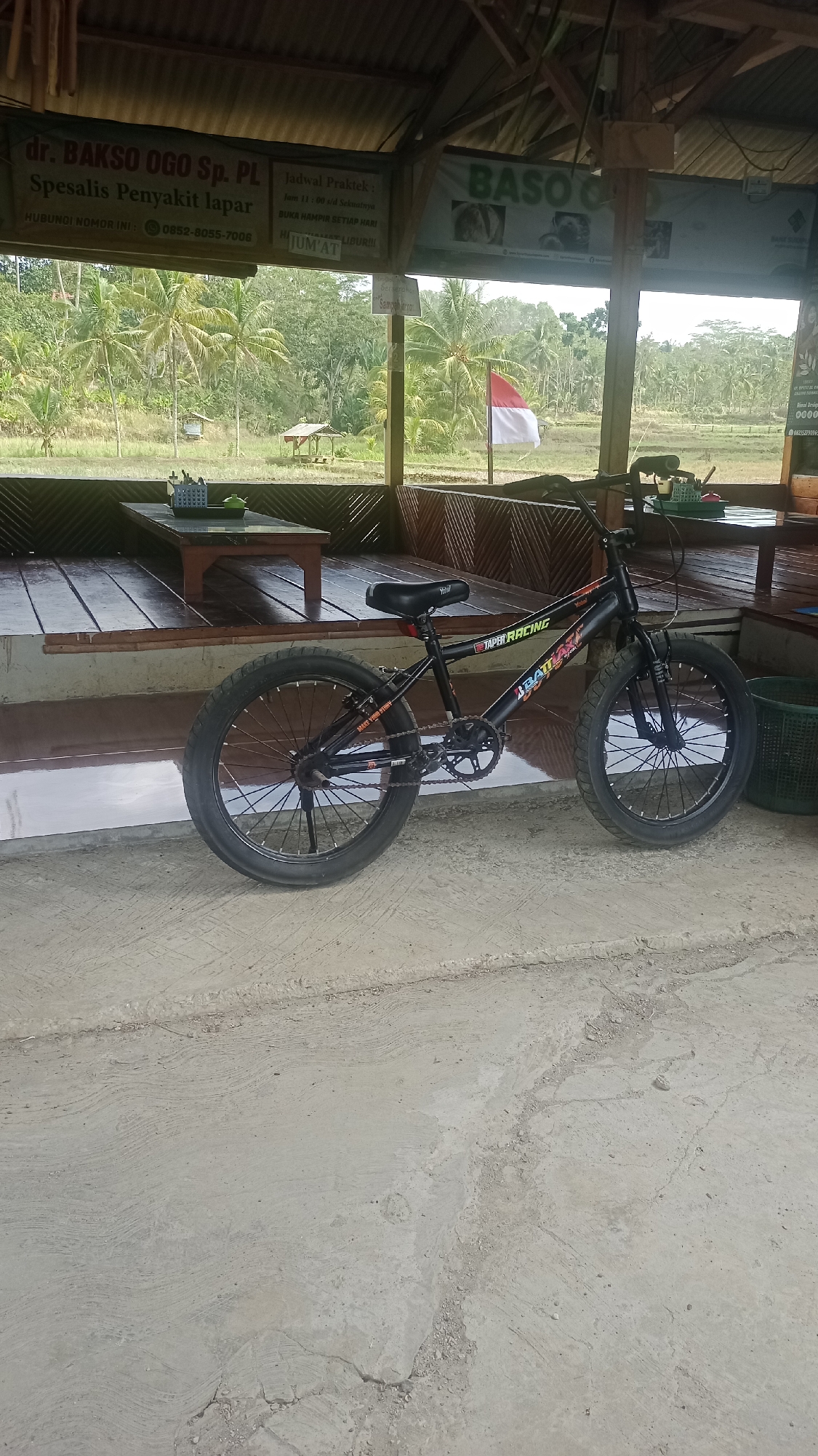 An outdoor, medium shot shows a black BMX bicycle parked on a concrete surface in front of a wooden structure. The bicycle is positioned to the right of the frame, facing towards the left. It has thick tires and a black seat. Behind the bicycle, a wooden platform is visible, with two low wooden tables and benches. Beyond the platform, a lush green landscape with palm trees and rice paddies unfolds. A small red and white Indonesian flag is seen flying on a pole in the distance. Several signs are affixed to the wooden structure. One sign, partially visible on the left, reads "dr. BAKSO OGO Sp. PL Spesialis Penyakit lapar". Another sign on the right has "BASO OGO" prominently displayed. The overall environment suggests a rural or semi-rural setting in Cibeber, Indonesia, likely a food stall or resting area. The lighting indicates daytime, with natural light illuminating the scene.