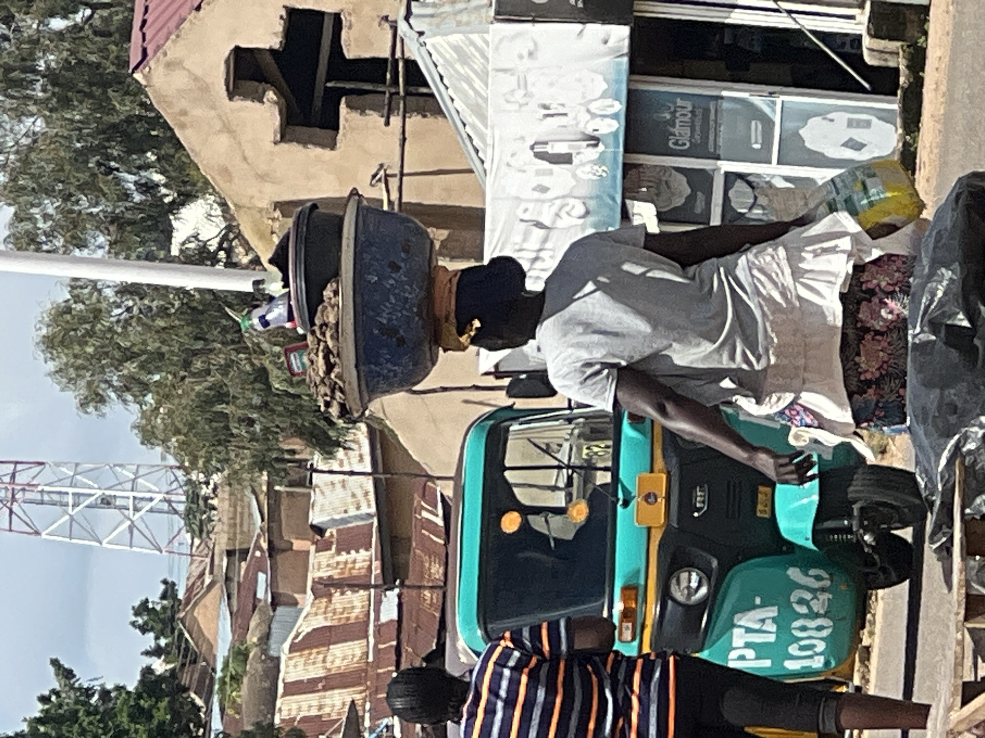 The image captures a street scene in Jos, Nigeria, under a bright, cloudy sky. In the foreground, a woman with dark skin and black hair is walking away from the camera, carrying a large blue basin filled with dark material on her head. She wears a light grey t-shirt and a white ruffled skirt over a floral-patterned fabric, and is holding a yellow and green plastic bag in her right hand. Beside her, to the left, is a teal and yellow auto-rickshaw with the license plate "PTA-10826". Another person, partially visible on the left, wears a horizontally striped orange and black t-shirt and dark pants.
In the background, residential buildings with corrugated metal roofs are visible. A tall, white lattice tower, likely a telecommunications mast, rises above the rooftops. Further in the background, there is a building with a red roof and a unique cross-shaped opening in its tan-colored facade. A shop with the name "Glamour" is partially visible to the right, featuring glass doors and windows displaying various items. The overall impression is a bustling urban environment with signs of commerce and daily life.