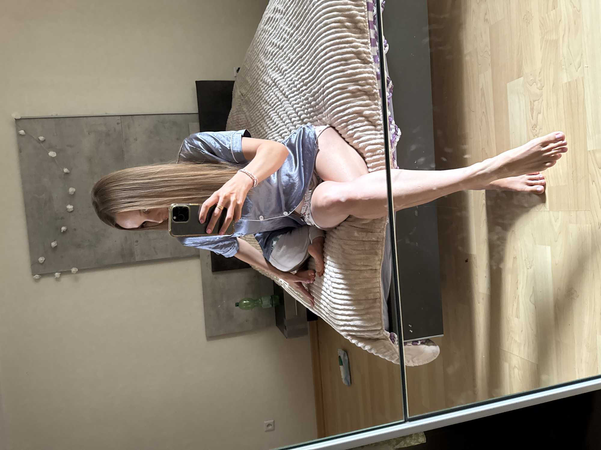 A young Caucasian woman with long blonde hair is sitting on the edge of a bed and taking a selfie with her phone. She is wearing a light blue pajama set. The bed has a textured beige comforter. To the left of the bed, a nightstand with a green water bottle and a framed picture hangs on the wall. The room has wooden flooring and a large mirror is in the foreground reflecting the scene. The overall setting appears to be a bedroom in a private residence, likely in Überherrn, Germany.
