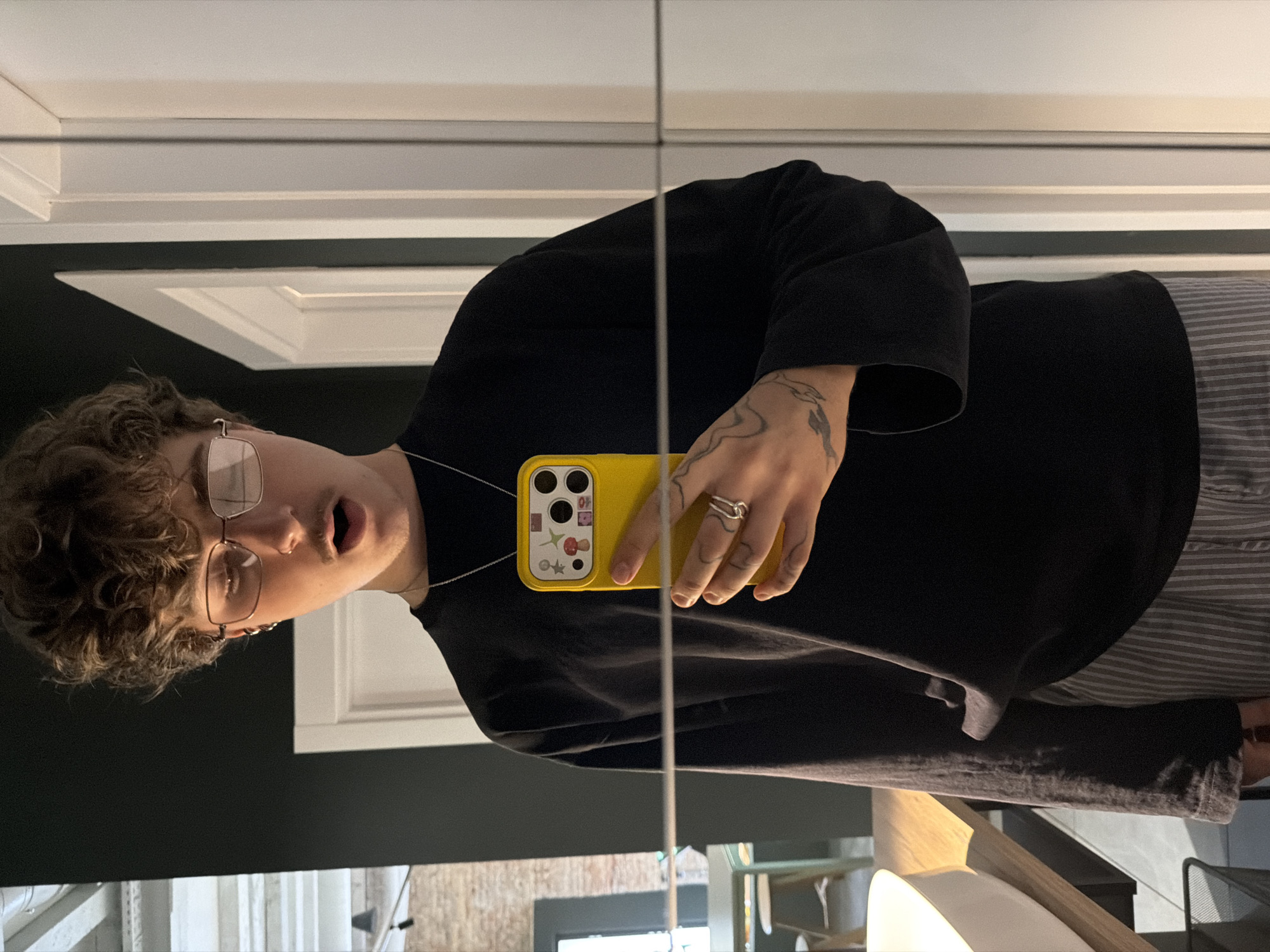 A person is standing in front of a mirror, taking a selfie. They have curly brown hair, are wearing glasses, and have their mouth open. They are wearing a black long-sleeved shirt and striped pants. They are holding a yellow phone in their right hand. The background shows a room with dark walls and white doors. The image is taken in a dimly lit interior. The reflection in the mirror shows a sink and counter on the left side of the frame, and a table and chairs in the background. There are no specific details in the image that would indicate the location of Belgrade, Serbia.