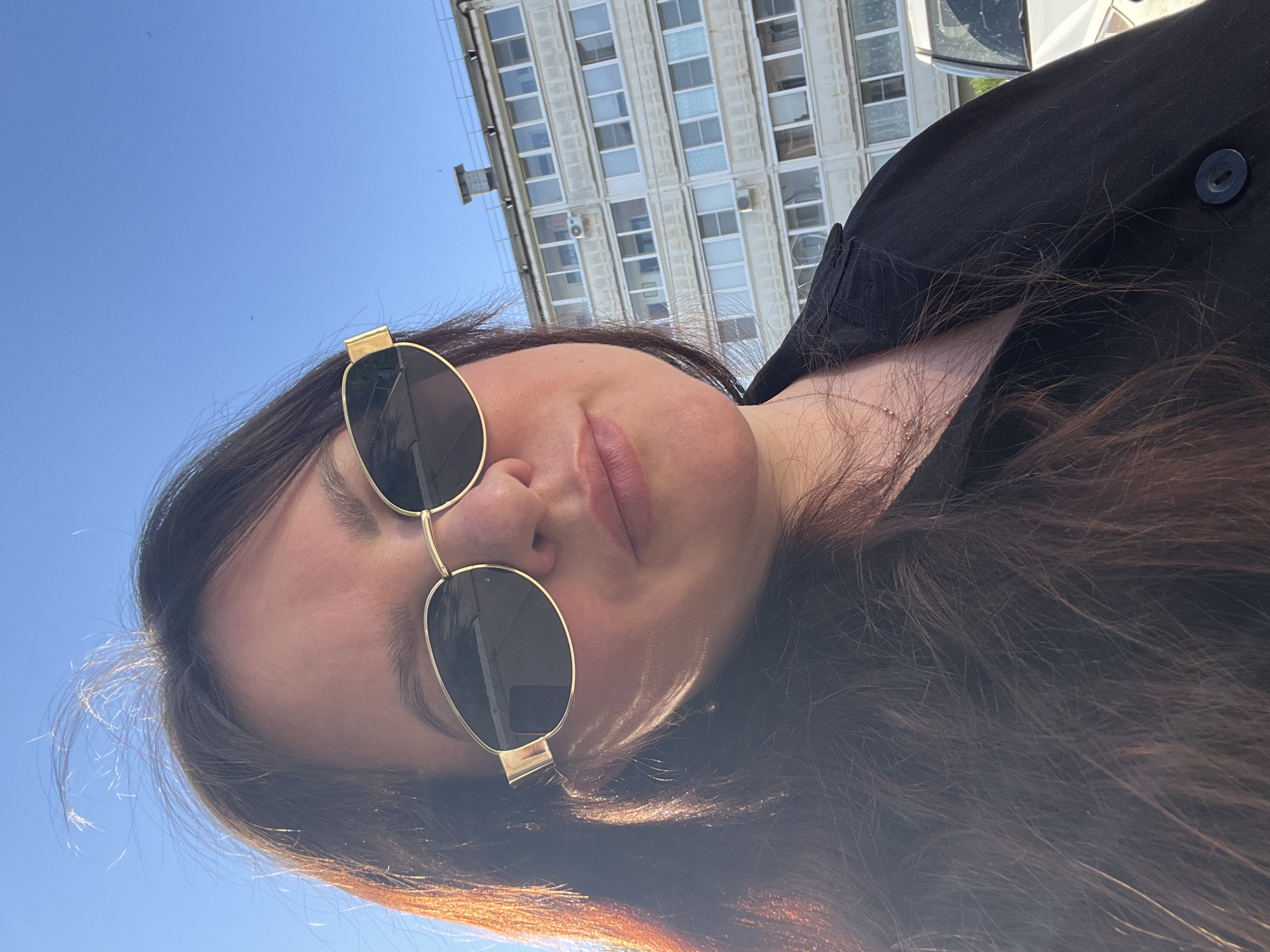 A woman wearing sunglasses poses for a photograph outdoors. In the background is a multi-story building with many windows. The woman has brown hair and is wearing a black jacket. The sky is blue. The city in which the photo was taken is not discernable based on the image alone.