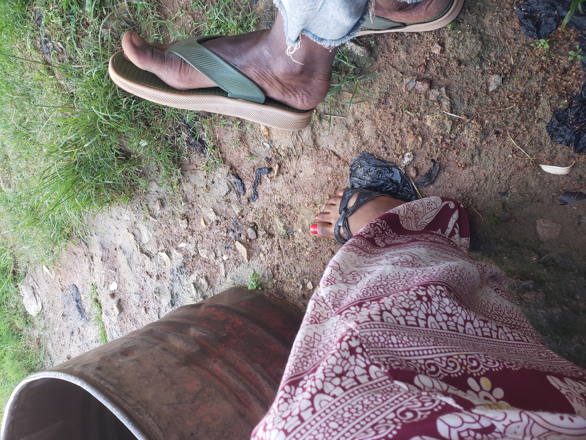 The image shows a close-up view of two people's legs and feet, situated outdoors on a dirt and grass ground. One person, on the left, is wearing a maroon and white patterned skirt and black sandals. Their toenails are painted red. The other person, on the right, is wearing blue denim jeans and green and beige flip-flops. They appear to be sitting or resting near a large, rusty barrel on the left side of the frame. The ground is a mix of soil, small stones, and sparse green grass, with some patches of wetness and dark debris. The overall scene suggests a casual, outdoor setting, possibly in a rural or less-developed urban area.