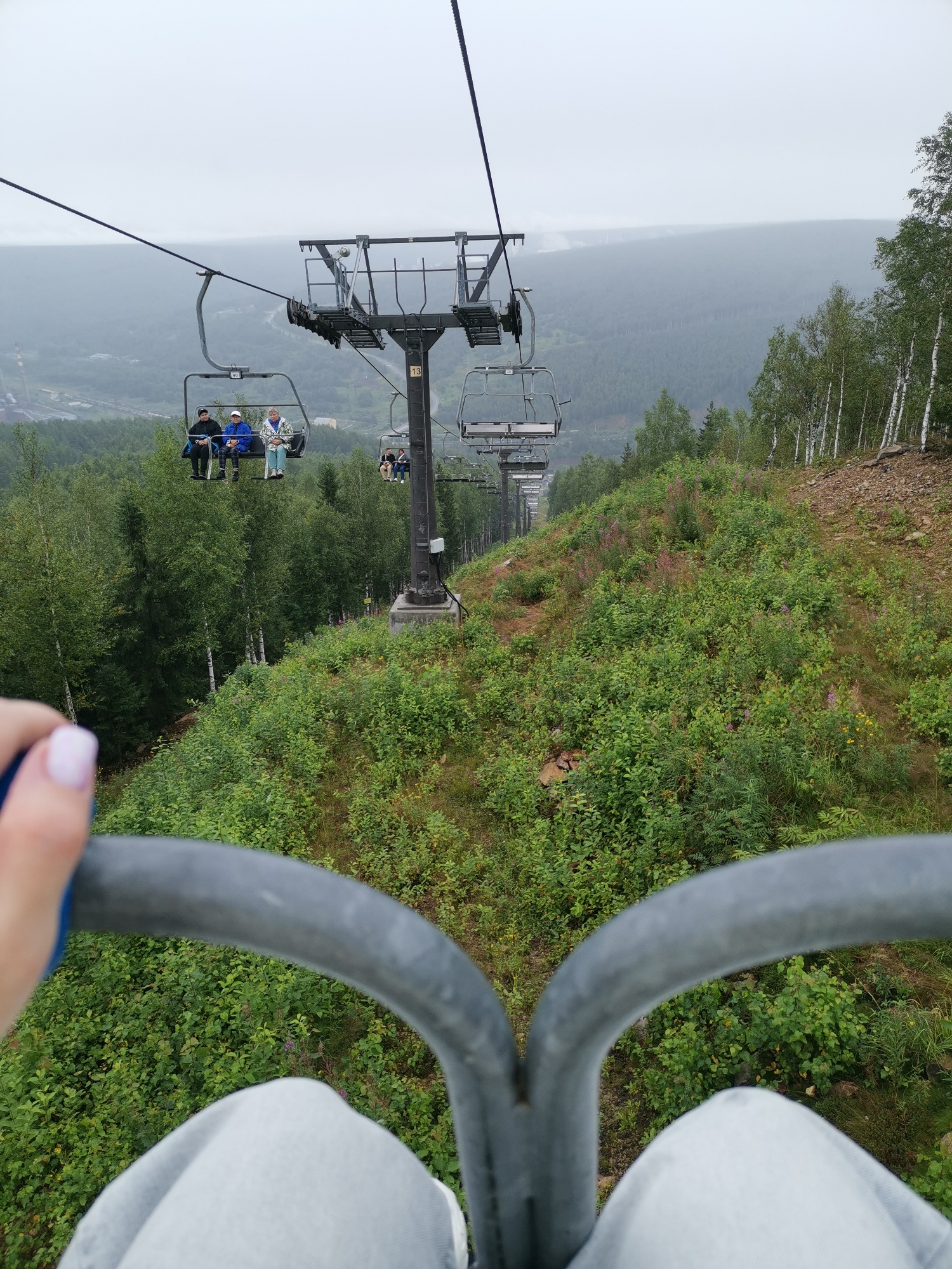 The image depicts a view from a ski lift in Gubakha, Russia, on an overcast day. A person's hand with light pink nail polish is visible in the foreground, gripping the grey metal bar of the ski lift. The ski lift is carrying several individuals, with three people in the immediate foreground and others further down the line. The chairs are suspended from a thick cable, which stretches across the frame. Below, a lush green hillside is covered in dense foliage and trees, with some birch trees interspersed among them. In the distance, a misty landscape of rolling hills and forests can be seen, with a faint indication of a road winding through the terrain. The overall atmosphere is serene and natural.