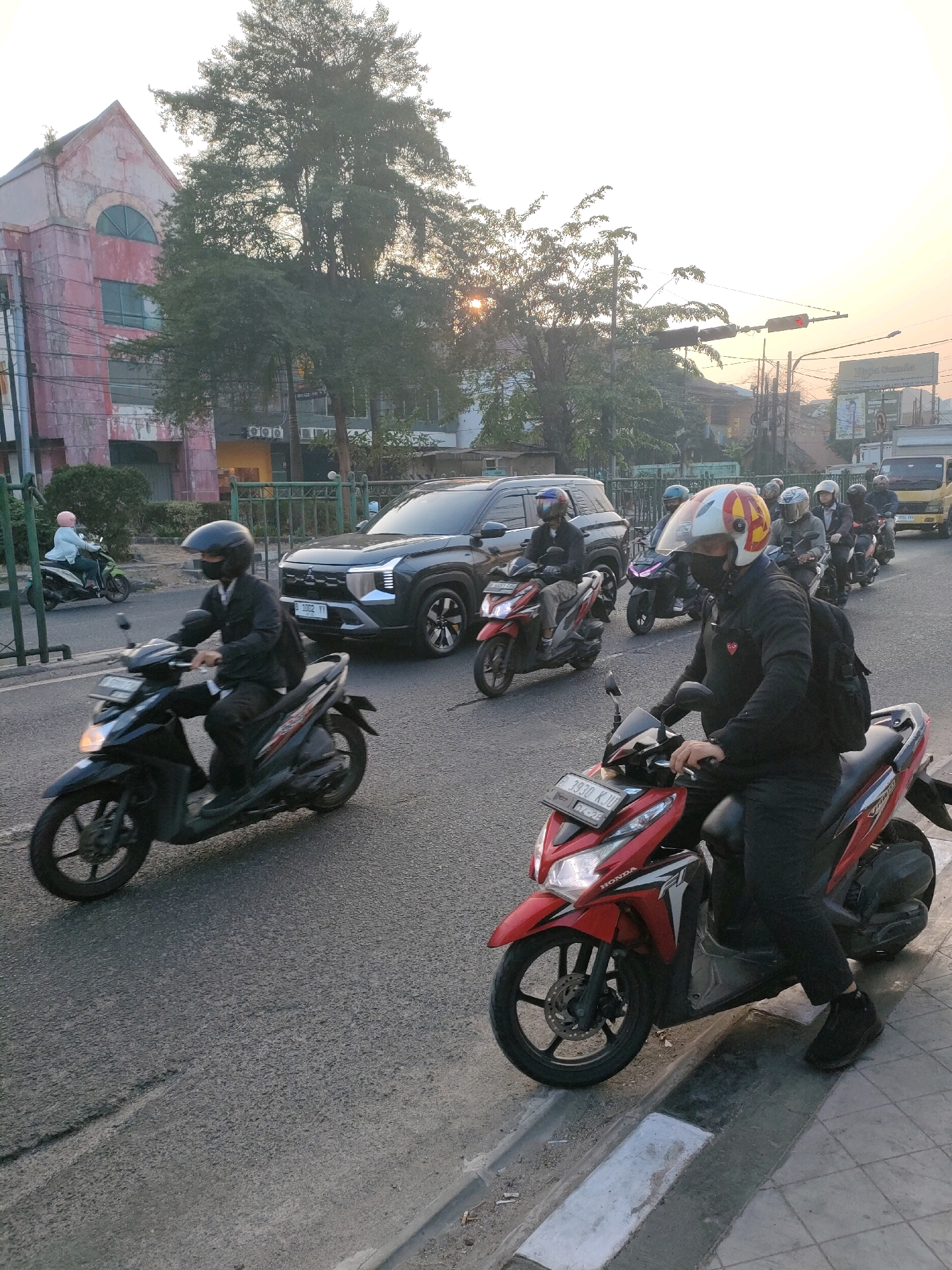 In the city of Bekasi, Indonesia, a street scene unfolds at what appears to be dusk, with the sun setting. A line of motorcycles and a dark SUV are captured on an asphalt road.
In the foreground, a man wearing a black jacket, black pants, and a black mask rides a red Honda scooter. He has a black backpack slung over his shoulders and a white helmet with red and yellow accents. The scooter's headlights are illuminated.
Behind him, another scooter, predominantly black, is ridden by a person in a black jacket and black helmet, also wearing a mask.
Further back, several other motorcycles and the dark SUV are visible. The SUV, a Mitsubishi Xpander, has its headlights on. Several riders on other motorcycles are also wearing helmets and masks.
To the left of the road, a pinkish-toned building with an arched window can be seen, partially obscured by trees. A green metal fence runs along the side of the road. In the distance, traffic lights are visible, with one signal displaying a red light. A yellow truck is parked further down the street. The sky exhibits a warm, hazy glow, indicative of the setting sun.