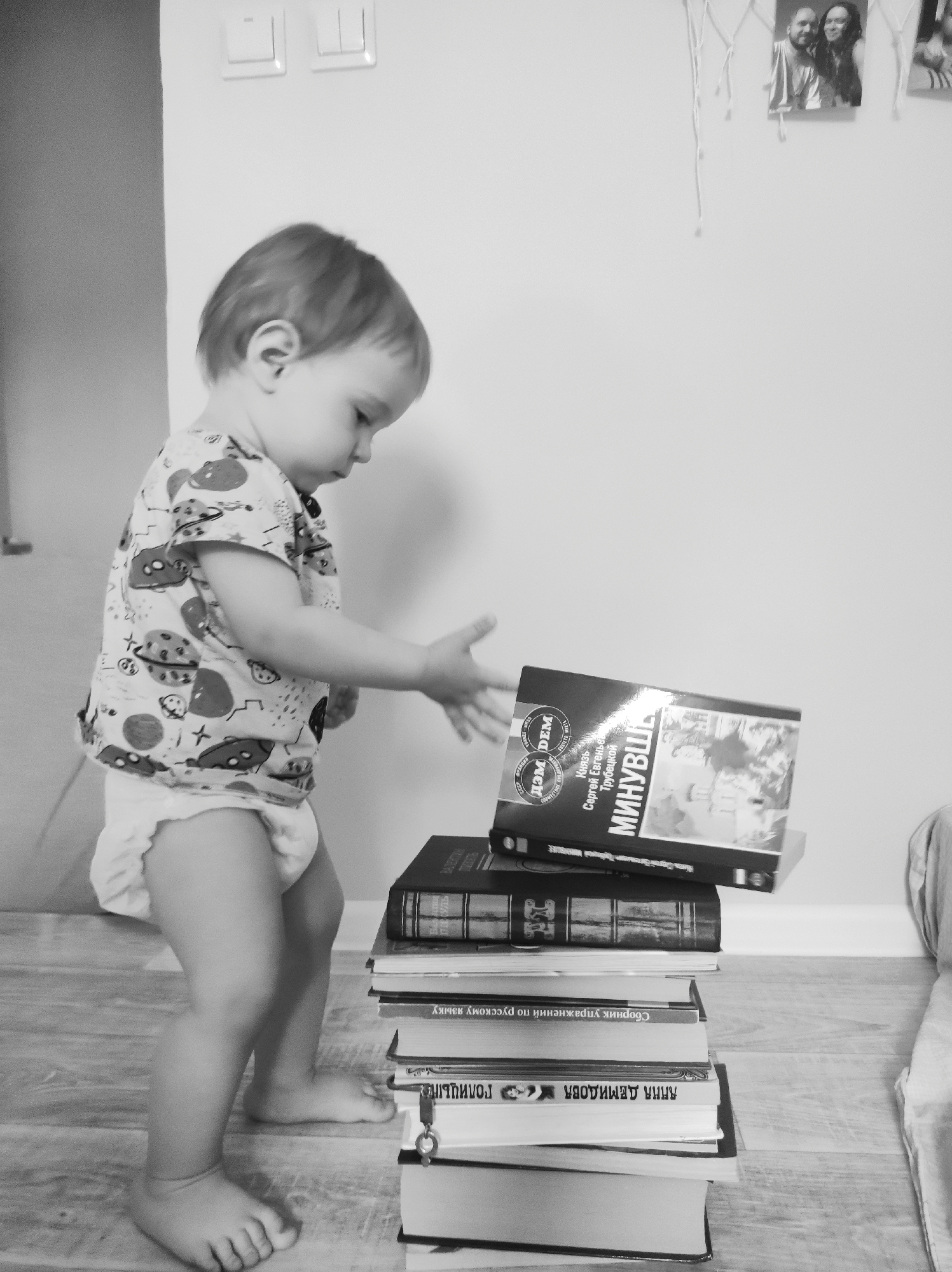 A grayscale, eye-level shot shows a baby attempting to grab a book from a stack of books. The baby is wearing a shirt with a pattern and a diaper. The baby's right arm is extended out to the books with the hand open, as if they are trying to grab a book. The stack of books is tall, and the topmost book is titled "Mинувшe" in Russian. The room has a light gray floor that appears to be made of wood. The wall behind the baby and books is white. On the upper-right of the shot, there are photographs hanging on the wall. There are light switches on the wall near the upper-left corner. The location is unknown, Unknown.