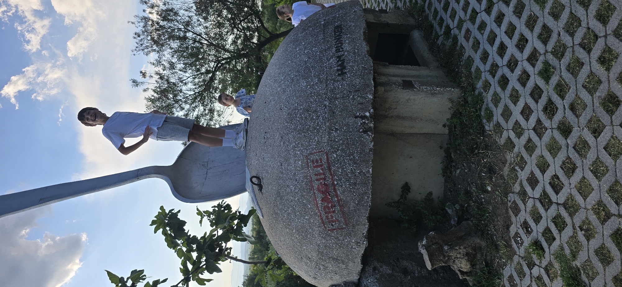 The photograph shows three people at the site of the monument known as the "Spoon Bunker," located in Blinisht, Albania. A teenager in a white t-shirt and gray shorts stands on a large metal spoon sculpture, part of the monument. A young girl in a blue floral dress is standing on top of a cement bunker that is also a part of the monument. A person is standing behind the bunker partially visible. The concrete bunker has the words "FRAGILE" and "HAN BUNKER" written on it. Paving stones with grass growing through them make up the ground. The sky is visible and there is a tree in the background.