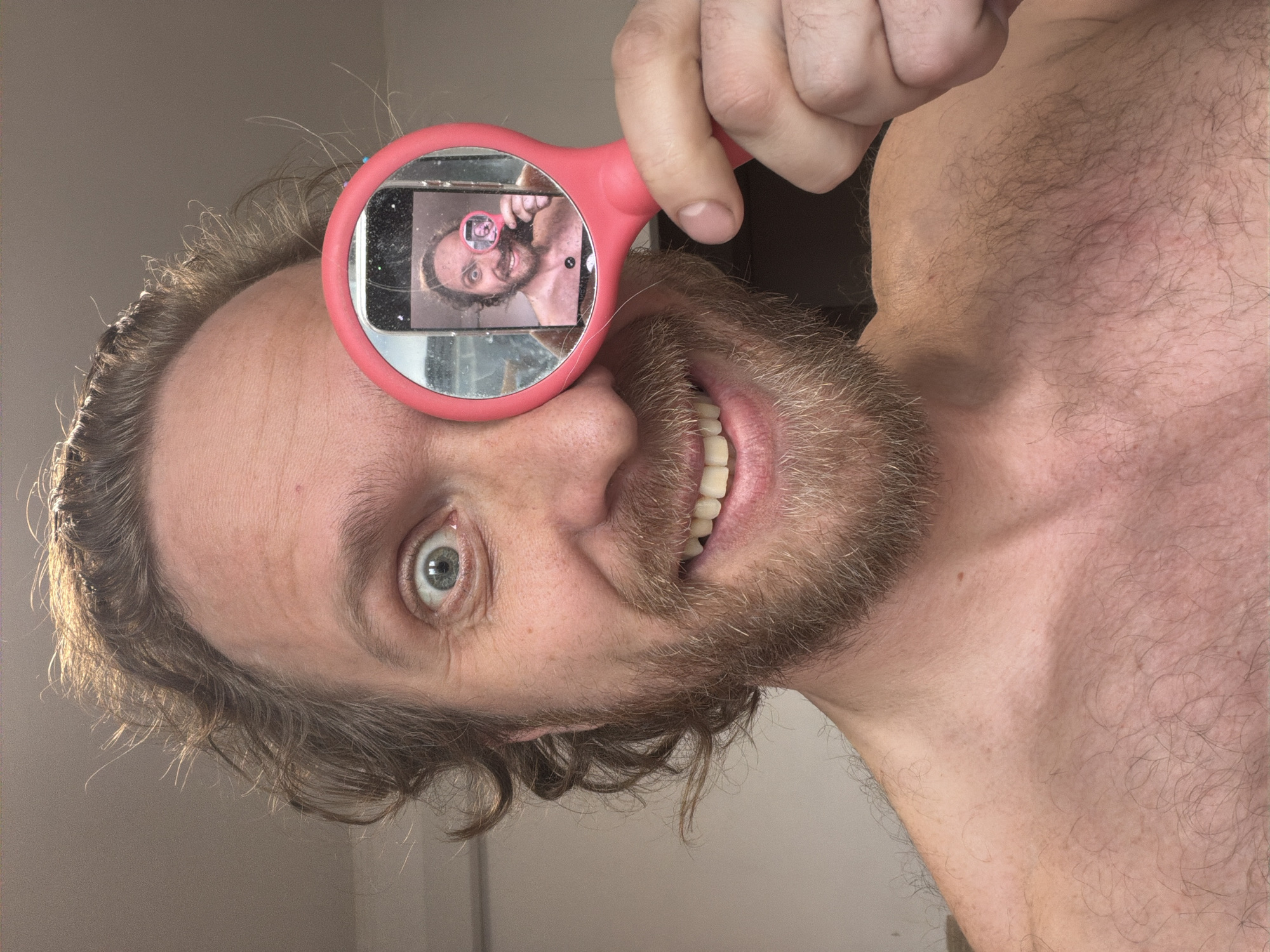 A man with shoulder-length brown hair and a beard is holding a pink-framed magnifying glass in front of his left eye. The magnifying glass reflects the image of a smartphone, which is taking a selfie of the man with the magnifying glass. The man is smiling, showing his teeth. The background is blurry, but appears to be a plain wall. The man's upper chest is visible, and he appears to be shirtless. There is no evidence or information indicating that this image was taken in Saint Petersburg, Russia.