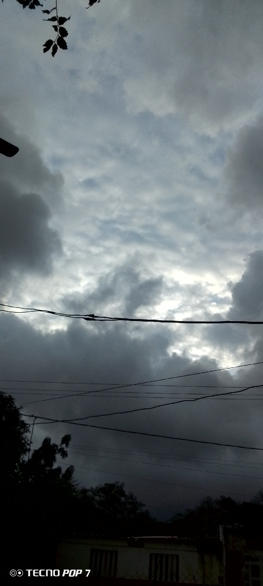 The image depicts an outdoor scene with a focus on the sky and the silhouettes of trees and man-made structures against it. The sky is overcast with dramatic, dark clouds, suggesting impending rain or a stormy weather event. Sunlight attempts to break through in patches, creating a dramatic contrast between light and shadow in the clouds.
In the foreground, several dark wires are strung across the frame, adding a sense of urban or suburban presence. Below the wires, the dark silhouettes of trees and the top of a building with barred windows are visible. The building appears to be made of light-colored material, possibly painted white or cream, and has a reddish-brown fence in front of it.
The image is captured in a way that emphasizes the atmospheric conditions. The time of day appears to be late afternoon or early evening, given the low light and the dramatic sky. The overall mood is somber and perhaps a little foreboding due to the dark clouds.
The text "© TECNO POP 7" is visible in the bottom left corner of the image, indicating the device used to capture the photograph and a copyright notice. There are no people visible in the image. The location context is Altagracia, Venezuela.