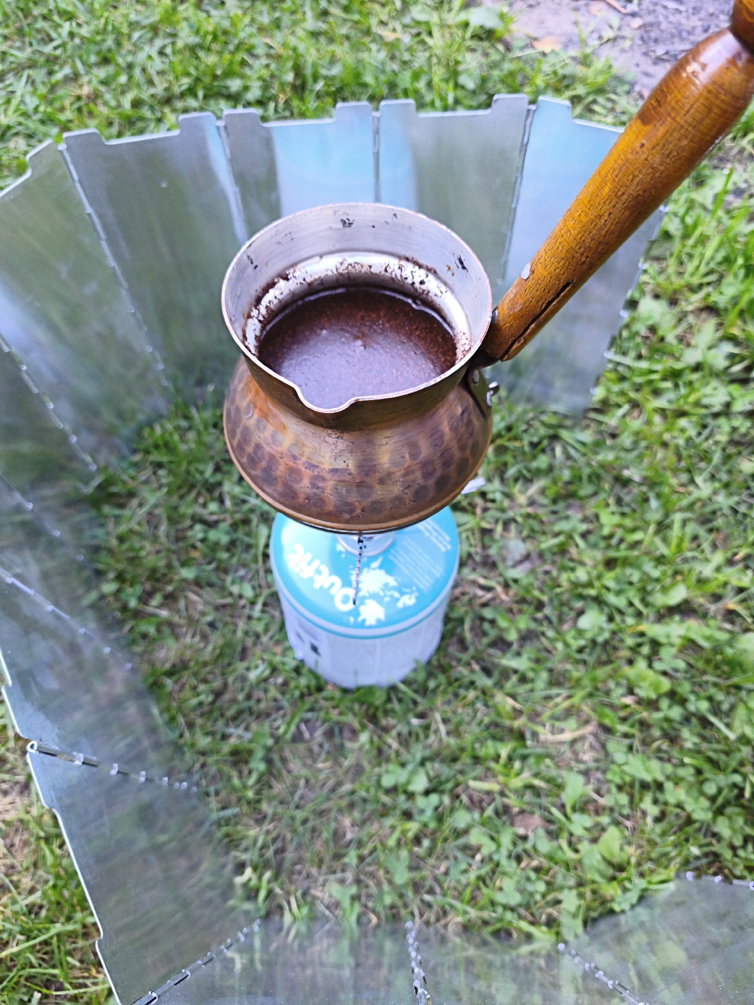 A close-up, high-angle outdoor shot captures a scene of coffee brewing. The main subject is a traditional copper cezve, filled with dark coffee, resting on a light blue gas canister. The cezve has a wooden handle, showing signs of wear and a crack. The brewing setup is enclosed by a metallic, segmented windscreen, common for camping or outdoor cooking. The background consists of lush green grass, suggesting an outdoor setting, possibly a park or a backyard, in Maardu, Estonia. The lighting indicates it is daytime, and the overall impression is one of a simple, rustic preparation of coffee in an outdoor environment.