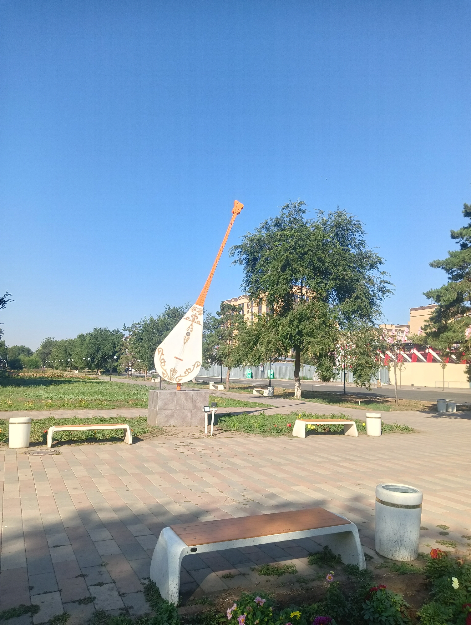 The image depicts a sunny day in a park located in the city of Aktobe, Kazakhstan. In the foreground, a paved walkway with interlocking colored bricks runs horizontally. Several modern-style benches with concrete bases and wooden seats are positioned along the walkway and in the grassy areas. Two concrete trash receptacles are visible.
Dominating the center of the image is a large, stylized sculpture of a dombra, a traditional Kazakh string instrument. The sculpture is white with intricate orange and gold decorative patterns on its body, and its orange neck extends upwards at an angle. It is mounted on a square, stone pedestal.
Behind the sculpture, a lush green park area features well-maintained grass, flower beds with colorful blooms, and numerous trees. A pathway winds through the park. In the background, apartment buildings with light-colored facades and some construction with red and white accents are visible. The sky is clear and blue. The overall scene conveys a sense of public space with decorative elements and recreational facilities.