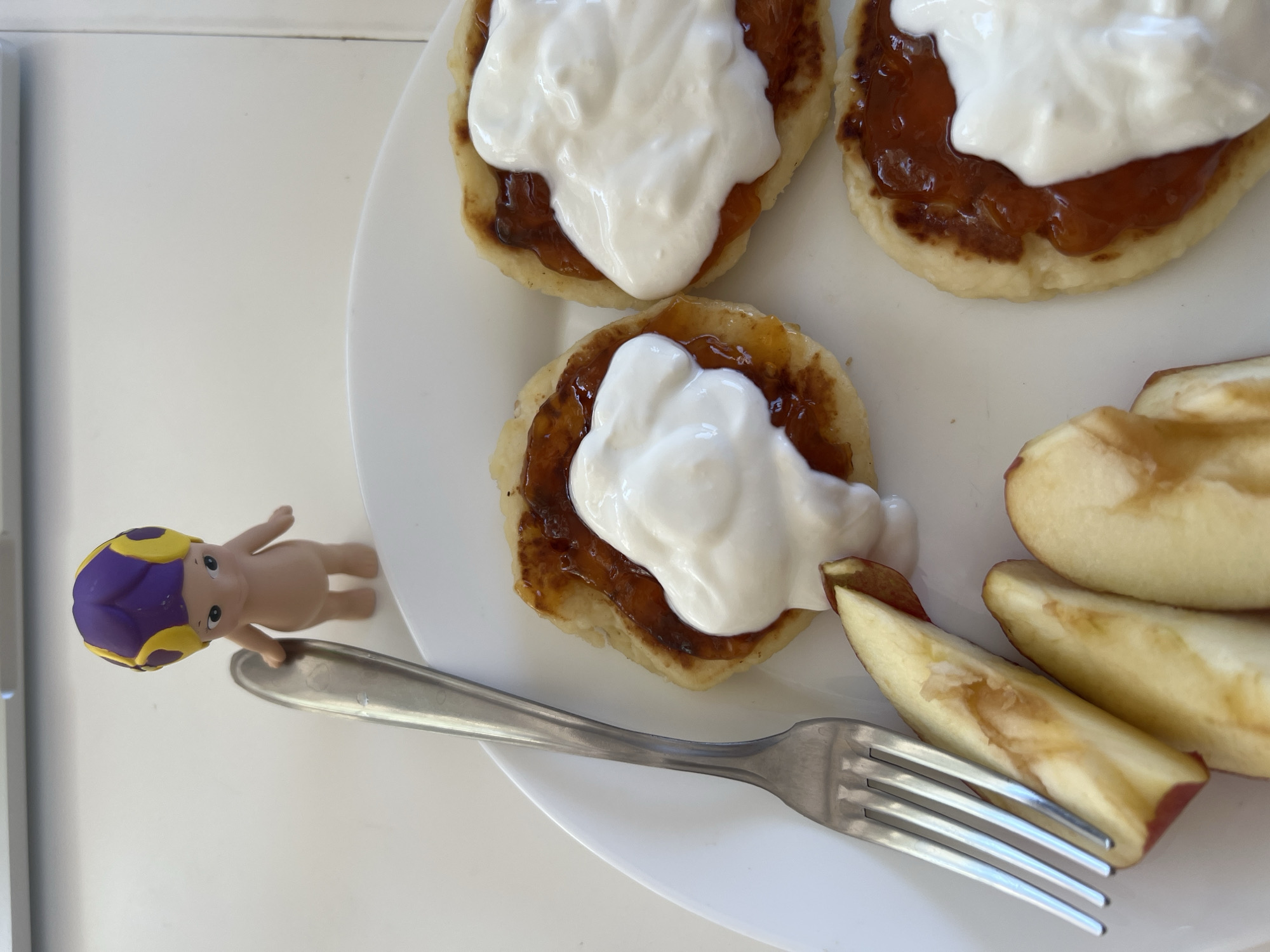 This close-up, overhead shot captures a simple meal presented on a white plate, set on a light-colored table, likely indoors. The main subjects are three small, round pancakes or fritters, each generously topped with an amber-colored jam or fruit preserve, and then crowned with a dollop of thick white cream, possibly sour cream or whipped cream.
Arranged to the right side of the plate are several slices of red-skinned apple, showing yellowish flesh and some light browning, indicating they were cut sometime prior to the photograph. A silver metal fork rests on the plate, with its tines partially obscured by one of the apple slices.
To the left of the plate, on the white table surface, stands a small, naked, baby-like figurine. This figurine features a prominent purple and yellow striped swimming cap, suggesting a playful or whimsical context to the scene. The lighting is bright and natural, indicating it is daytime. No text is visible in the image. The overall impression is that of a casual and pleasant meal setup.