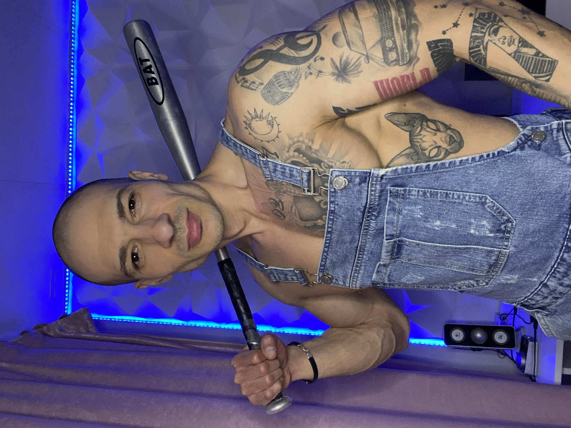 The image displays a bald adult male in an indoor setting in Pleven, Bulgaria. He is presented from the waist up, facing the camera with a neutral expression. The man wears unfastened blue denim overalls, exposing his heavily tattooed chest and torso. He holds a silver-colored baseball bat, marked "BAT" on its barrel with a black taped grip, over his right shoulder. A black bracelet is present on his left wrist. Visible tattoos include a sun and moon, a microphone, musical notes, a palm tree, a car, and an Egyptian pharaoh's head on his right arm, and a cat's head on his left torso. The background features a wall section with geometric, three-dimensional panels illuminated by blue LED strip lights. A light-colored curtain hangs on the left. A black speaker, identified by a "Z4" logo and three circular components, rests on a white surface below the subject's left arm, accompanied by visible wires. The scene is primarily bathed in a blue ambient light.