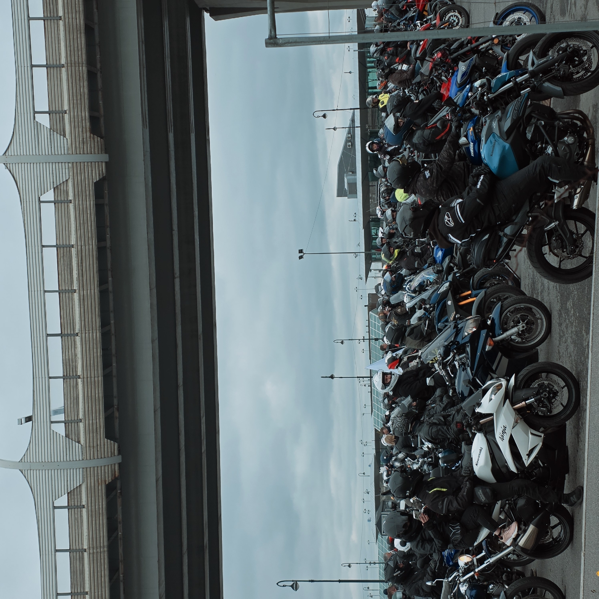 The image shows a large group of motorcycles parked in a row. Many motorcyclists are sitting on them. The scene appears to be an urban setting, likely in Saint Petersburg, Russia. A structure is visible in the image that appears to be the Saint Petersburg stadium that sits on Krestovsky Island, which is near a bridge. The sky is overcast, suggesting a cloudy day.