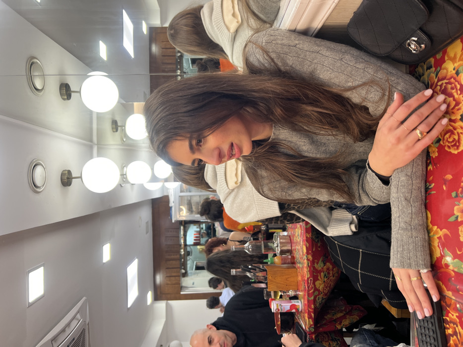 A woman with long brown hair wears a gray sweater and sits at a table covered in a red and yellow floral pattern. A black purse rests on the table. She wears rings on her fingers and looks down. The background shows a restaurant interior with other patrons, a mirror, and a row of round light fixtures.