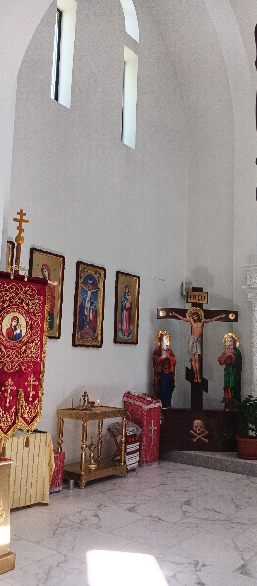 The image shows the interior of an Orthodox church. The scene is dominated by religious iconography and furnishings. On the left, a large red banner with gold embroidery hangs, featuring an image of the Virgin Mary and child. To the right of the banner are three framed religious icons depicting scenes from the Bible. In the center of the image, a golden altar stands with lit candles. Further to the right, a large wooden crucifix with Jesus is prominently displayed. Two smaller religious statues are positioned on either side of the crucifix, and a skull and crossbones symbol is visible at the base of the crucifix. The walls are white, and the floor is tiled with a marble pattern. Two tall, narrow windows are visible in the upper portion of the image. This scene is consistent with the interior of the Resurrection of Christ Cathedral in Smila, Ukraine. There are no people visible in the image.