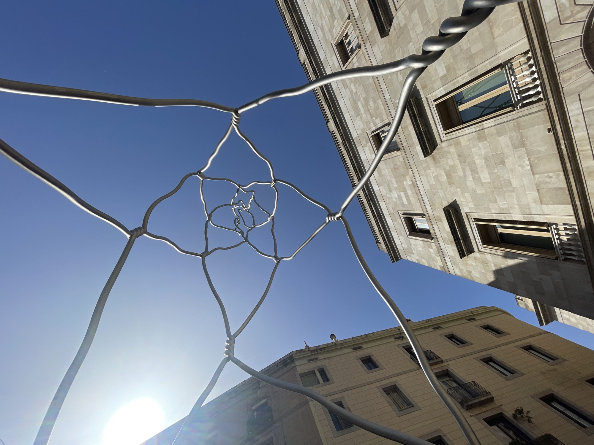 The photograph, taken from a low-angle perspective, captures a sunny day in Barcelona, Spain, with a clear blue sky. In the foreground, a metallic sculpture made of intertwined wire forms an intricate, spiraling pattern. The sculpture appears to be an abstract representation, possibly a fractal, with its delicate strands creating a sense of depth and complexity.
Behind the sculpture, two buildings are visible. The building on the left is lighter in color, possibly cream or pale yellow, and features multiple windows with balconies. The building on the right is constructed from stone blocks and exhibits architectural details like cornices and moldings. It also has windows with balconies and wooden shutters. The sun is bright, casting a lens flare in the upper left portion of the image.
The scene is devoid of people, and the focus is on the architectural elements and the striking metallic sculpture against the backdrop of the city. The composition suggests an urban setting, likely a public space or plaza in Barcelona, where art and architecture converge. The spiraling form of the sculpture draws the viewer's eye upwards towards the sky, creating a dynamic contrast with the static nature of the buildings.