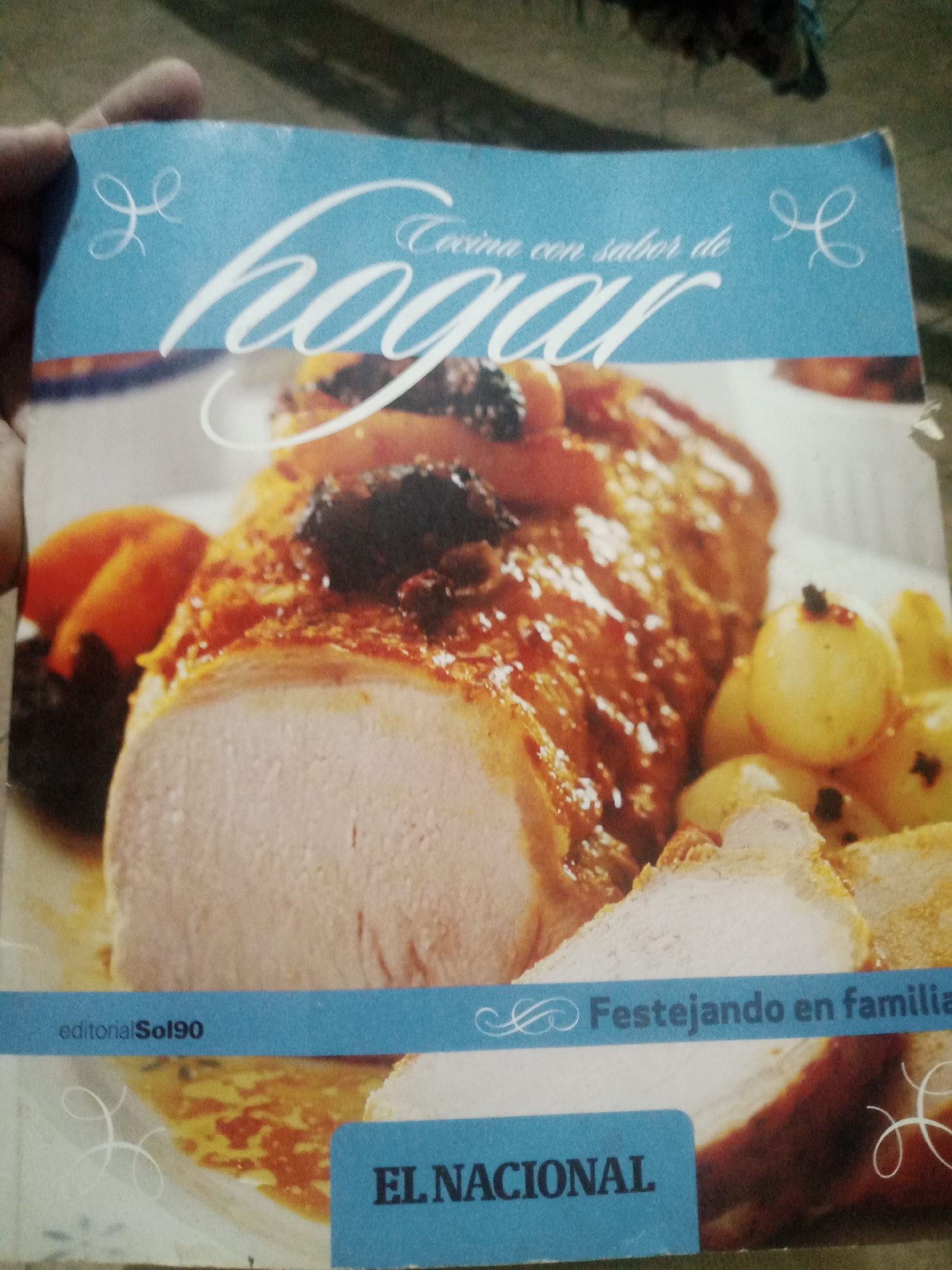The image shows a close-up of a cookbook cover featuring a roasted meat dish with what appears to be prunes and small onions. The cookbook has a blue banner across the top with the title "hogar" in large script, preceded by "Cocina con sabor de". There are decorative flourishes on either side of the title. Below the food, another blue banner reads "editorialSol90" on the left and "Festejando en familia" on the right, with a decorative flourish. A further blue section at the bottom has the words "EL NACIONAL" in large, bold capital letters. The cookbook is being held by a hand on the left side, with the fingers partially visible. The background is dark and out of focus. The image is an indoor photograph of a cookbook. There is no information in the image to suggest a location in Santa Ana, Venezuela.