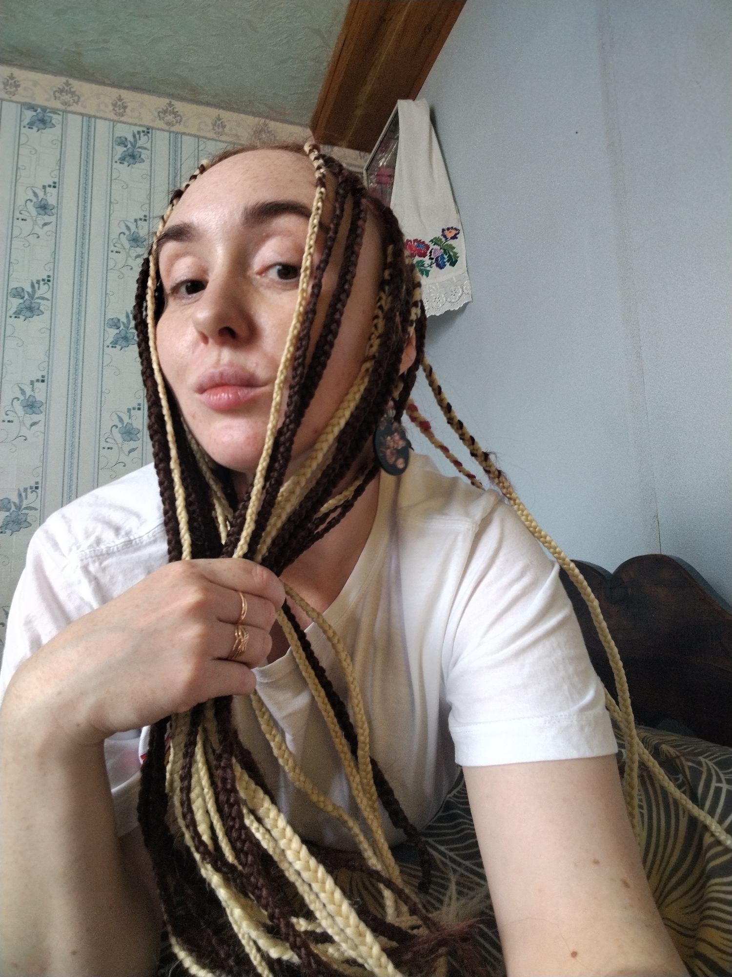 A person with light skin and freckles is indoors. They have dark brown and blonde braids. The person is wearing a white t-shirt. They are holding a bunch of the braids in their left hand, which has two gold rings on the ring finger. The background includes floral wallpaper, a light blue wall, and a wooden beam on the ceiling. A decorative embroidered towel hangs on the light blue wall. The location is inferred as being Яган, Russia from the query.