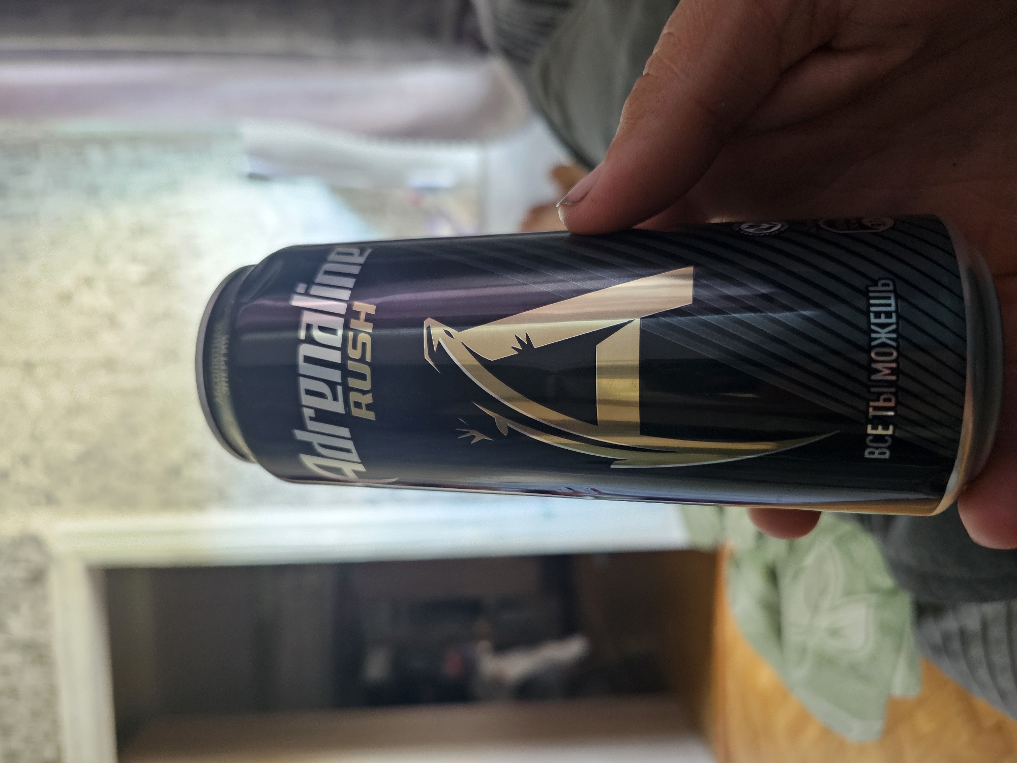 In Yelan, Russia, a close-up shot focuses on a hand holding a dark blue can of Adrenaline Rush energy drink. The can features the brand name in white capital letters with a stylized gold 'A' and a gecko graphic. The text "ВСЕ ТЫ МОЖЕШЬ" is visible at the bottom of the can. The background is blurred, showing a doorway with a light-colored wall and a glimpse of what appears to be a room with furniture and natural light filtering through a window. The lighting suggests it is daytime.