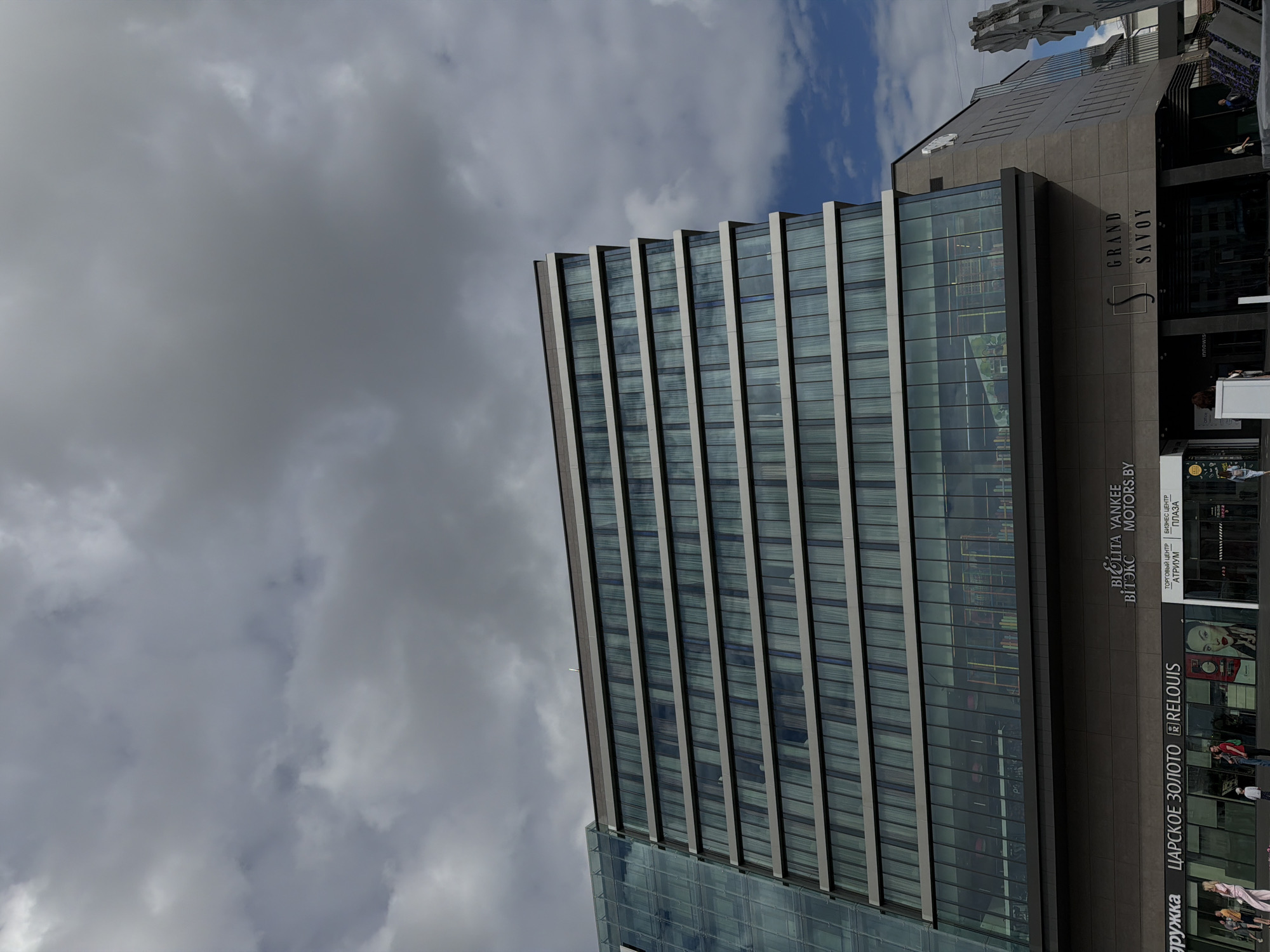 This image captures the Grand Savoy building in Mahilyow, Belarus, under a cloudy sky. The building features a modern architectural design with extensive glass facades and horizontal banding. The lower section of the building houses several businesses, including "ЦАРСКОЕ ЗОЛОТО" (Tsarskoye Zoloto), "RELOUIS," and "Grand Savoy." The sky is filled with gray and white clouds, with patches of blue sky visible. Several people are present in front of the building, walking and interacting. One person is standing in front of a kiosk displaying a graphic of a woman's face. The scene is illuminated by natural daylight.