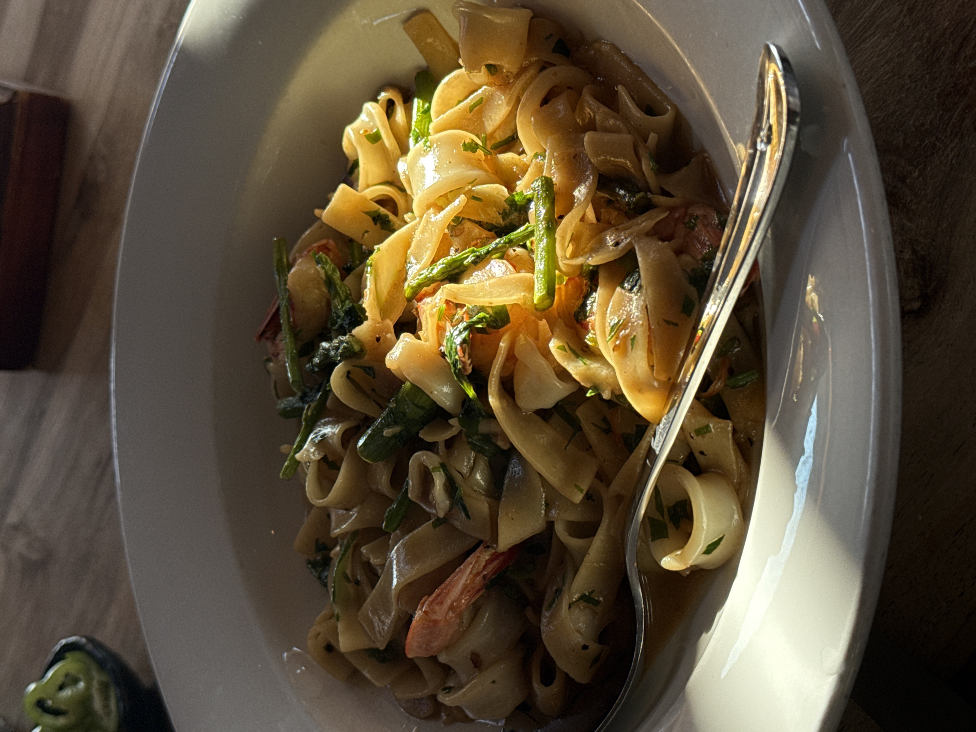 This is a close-up, eye-level shot of a pasta dish served in a white ceramic bowl. The pasta is wide and flat, resembling fettuccine, and appears to be tossed with a light sauce. Visible ingredients include chopped green herbs, pieces of asparagus, and what appear to be shrimp and calamari rings. A fork rests on the rim of the bowl, partially submerged in the pasta. The lighting suggests natural sunlight casting a strong shadow across the dish and the table it sits on. In the upper left corner, a small dark bowl contains sliced green peppers, likely a condiment or garnish. The table surface is made of wood. The overall scene is suggestive of a meal in a restaurant setting, possibly in Tel-Aviv, Israel, given the context of the prompt.