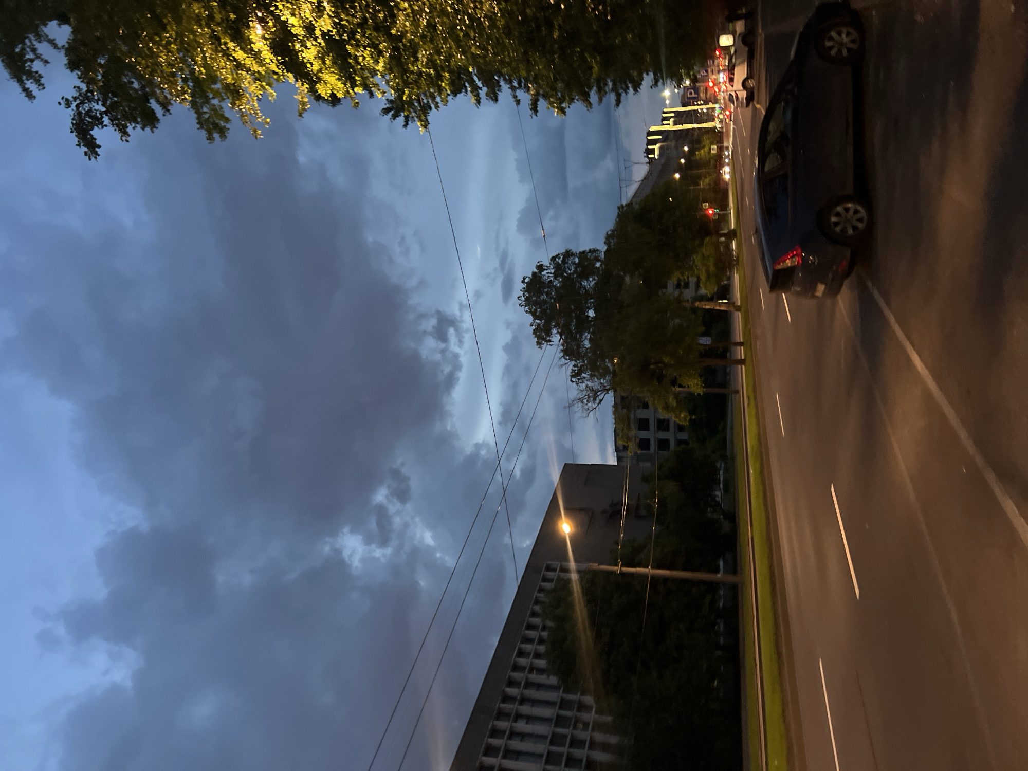 The image captures an urban street scene at dusk or night in Saint Petersburg, Russia, likely along Moskovsky Prospekt, looking south. A wide, multi-lane asphalt road with visible white lane markings extends into the distance, illuminated by warm-toned streetlights.
In the immediate foreground on the right, a dark-colored hatchback vehicle, possibly a Toyota Prius, is parked partially on a striped parking area and partially on the curb. Further along the right side of the street, trees with dense foliage are brightly illuminated by streetlights, causing their leaves to appear a vivid yellowish-green. In the midground, other vehicles are visible on the road, including a white car, and traffic signals display red lights. A modern building in the distance on the right features prominent vertical strips of yellow illumination on its facade.
On the left side of the road, a large, grey, multi-story building with a distinctive facade characterized by numerous horizontal window bands is partially obscured by trees. A brightly glowing streetlamp in the midground on the left emits a strong lens flare. Overhead wires, indicative of a trolleybus or tram route, crisscross the upper portion of the frame. The sky is heavily overcast with dark grey clouds, though a narrow, lighter blue-grey strip is visible towards the horizon, suggesting ambient light despite the heavy cloud cover. No individuals are discernible within the frame.