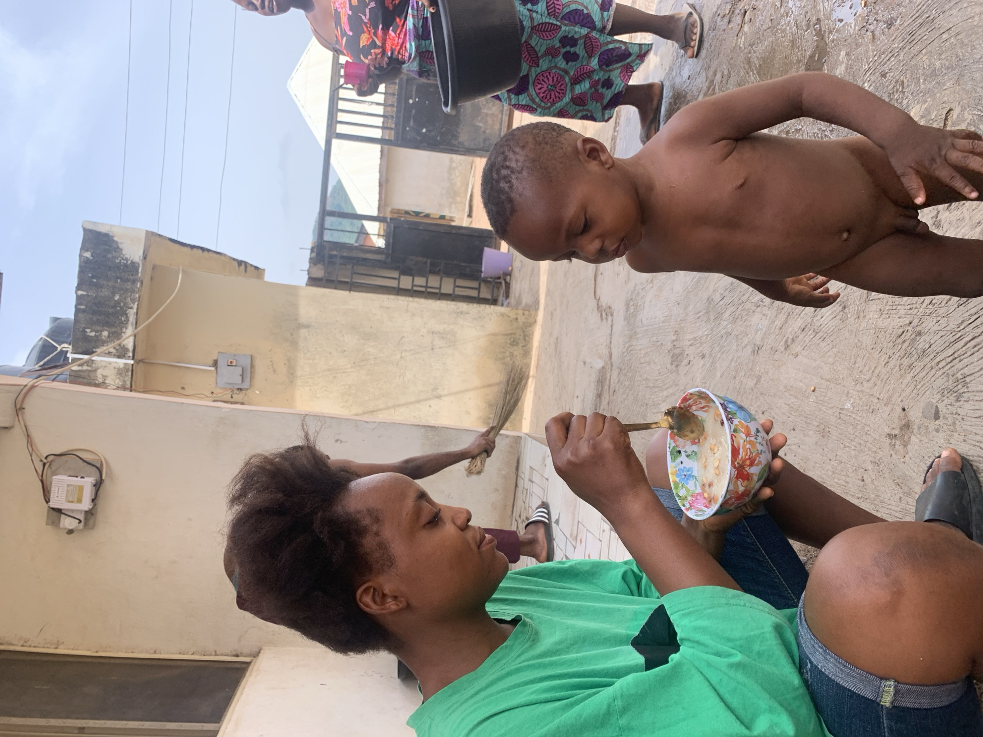 In a sunlit outdoor space in Lokoja, Nigeria, a dark-skinned child stands naked on a concrete surface. A woman with dark hair, wearing a green shirt and jeans, sits cross-legged, holding a floral-patterned bowl and spoon, seemingly offering the child food. Another adult, partially visible, holds a broom in the background. A woman with a floral dress carries a black bucket. Buildings and a metal gate form the backdrop. Electrical wires and a switch box are affixed to the building wall.