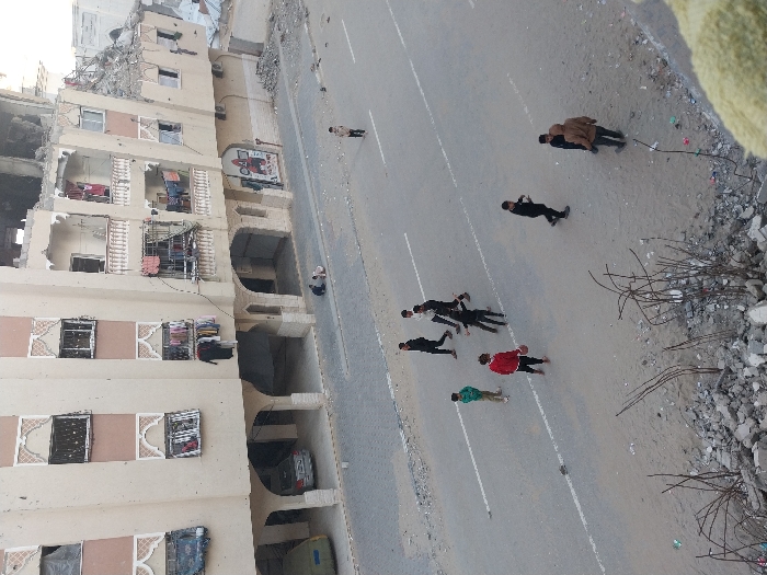 A damaged building stands beside a road where several people lie face-down. Some are in dark clothing, while others wear brighter colors like red and green. One person is being carried by another. Debris is scattered near the road, and a few individuals stand or walk on the roadside. The overall scene suggests a location impacted by conflict or disaster.