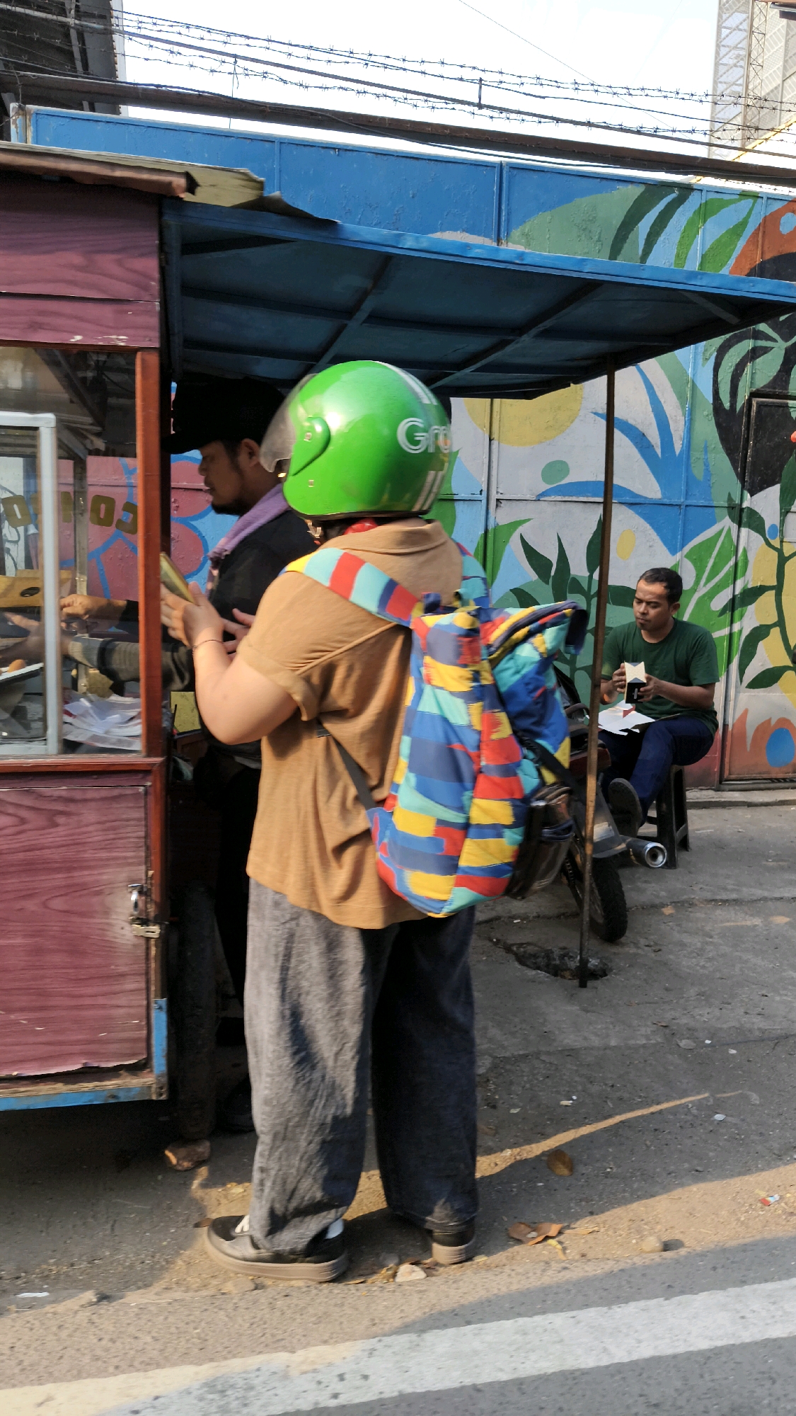 The image depicts an outdoor scene in South Jakarta, Indonesia, likely on a street. In the foreground, a person wearing a green motorcycle helmet with "Grab" branding and a colorful, geometric-patterned backpack is standing next to a food cart. The person is wearing a brown t-shirt and dark grey trousers. The food cart has a wooden exterior and a blue awning. Behind the food cart, another person in a black shirt and a purple scarf is attending to the cart. To the right, a man in a green t-shirt and dark blue jeans is seated on a black stool next to a motorcycle, looking down at a small object in his hands. The background features a vibrant mural with green and yellow abstract designs on a wall. Barbed wire is visible along the top of the image. The ground is paved and shows signs of wear and tear, with shadows cast by the sun.