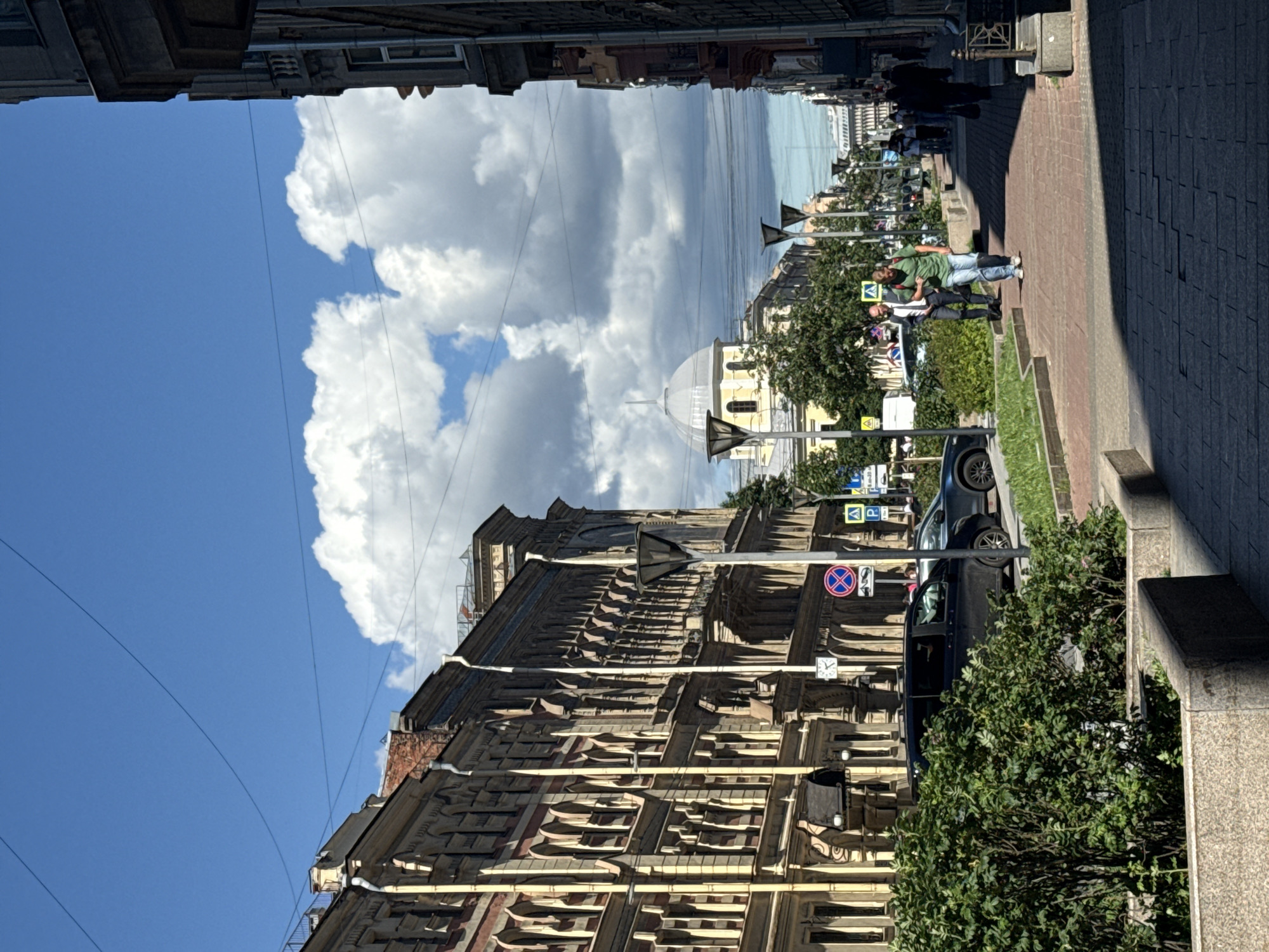 The image depicts a street scene in Saint Petersburg, Russia, on a sunny day with scattered clouds. A wide street is lined with ornate, historic buildings on the left, characterized by their multi-story facades, arched windows, and decorative architectural details. A streetlamp is visible in front of one of these buildings. On the right side of the street, a pedestrian walkway paved with red brick is visible, with several people walking along it. A man in a green shirt and jeans walks towards the camera, while another man in a suit walks in the opposite direction. A dark-colored minivan and a light-colored car are parked on the street on the left. Further down the street, a light-yellow domed building with a spire is visible, likely a church or government building. Green trees and bushes line the sidewalk. Overhead, telephone wires crisscross the blue sky. The overall impression is of a bustling urban environment with a blend of historical architecture and modern life.