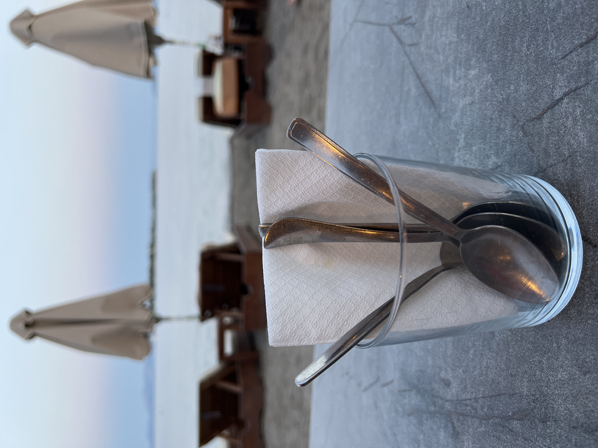 A close-up shot focuses on a clear glass filled with several spoons and a folded white napkin. The glass sits on a textured gray surface, likely a table. In the background, out of focus, is a sandy beach scene. Two closed beach umbrellas are visible, along with lounge chairs and the calm sea under a hazy twilight sky. The location is identified as Eretria Municipal Unit, Greece.