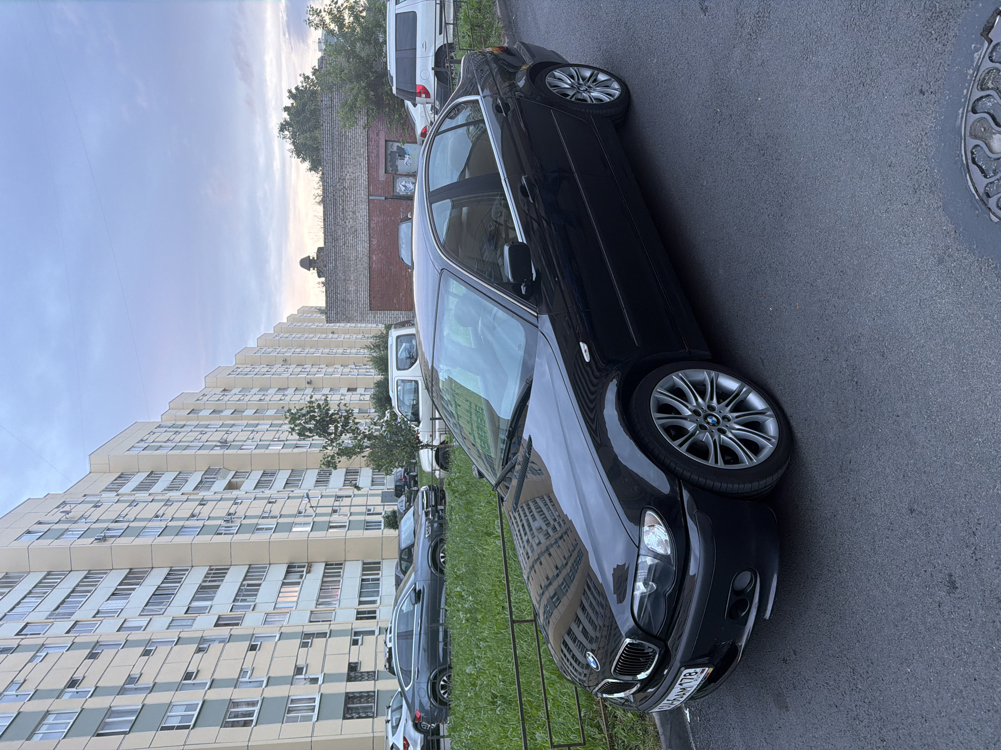 A black BMW sedan is parked on an asphalt street in front of a tall, multi-story apartment building. The building is light beige with light blue vertical accents and numerous windows. To the right of the apartment building is a low brick structure with a metal roof. Several other vehicles are visible in the background, including a dark gray sedan, a white van, and a white SUV. A patch of green grass with a metal fence separates the street from the apartment building. The sky is overcast with pale blue and gray clouds. The image appears to have been taken during the day.