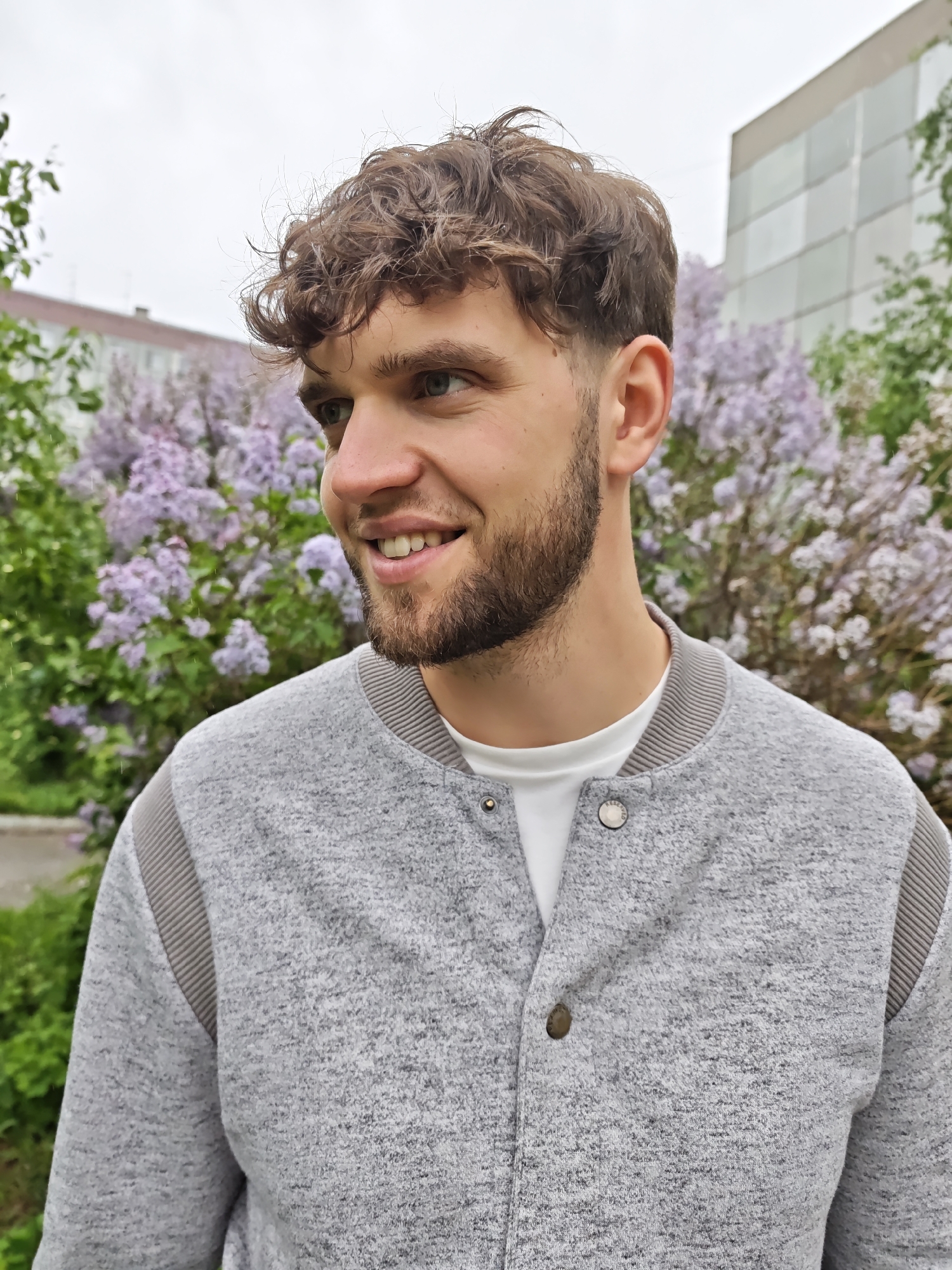 The image is a medium shot of a young man with curly brown hair and a short beard, smiling and looking to the left. He is wearing a light gray jacket over a white T-shirt. The jacket has gray ribbed detailing on the collar, cuffs, and shoulders. In the background, there are lilac bushes in full bloom, and glimpses of buildings can be seen, suggesting an urban setting. Based on the image data, the location is Novosibirsk, Russia. The scene is outdoors, likely in a park or garden area.