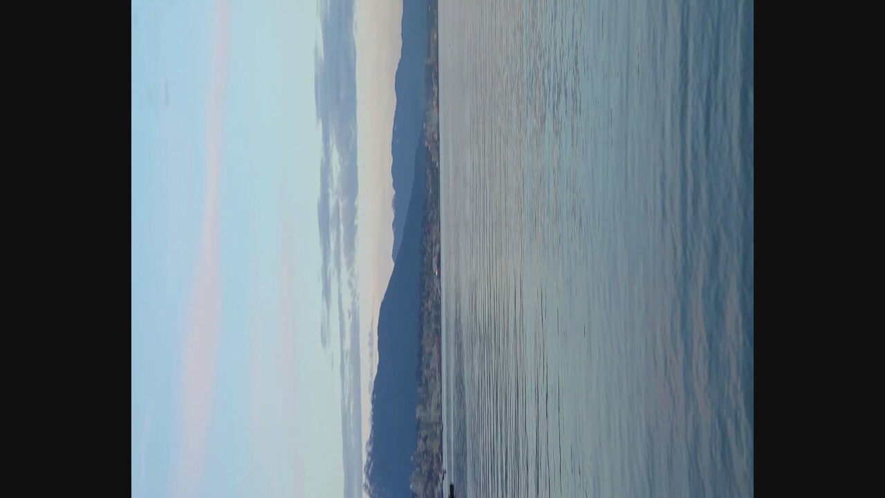 The image shows a wide view of the water in Vancouver, Canada, with mountains in the background. The sky is light blue with scattered pink and gray clouds. The water is calm with small ripples reflecting the sky. In the distance, the Vancouver skyline is visible along the waterfront, with buildings nestled against the backdrop of dark, forested mountains. The overall scene suggests a peaceful, natural setting within an urban environment, likely captured during the early morning or late evening when the light is soft.