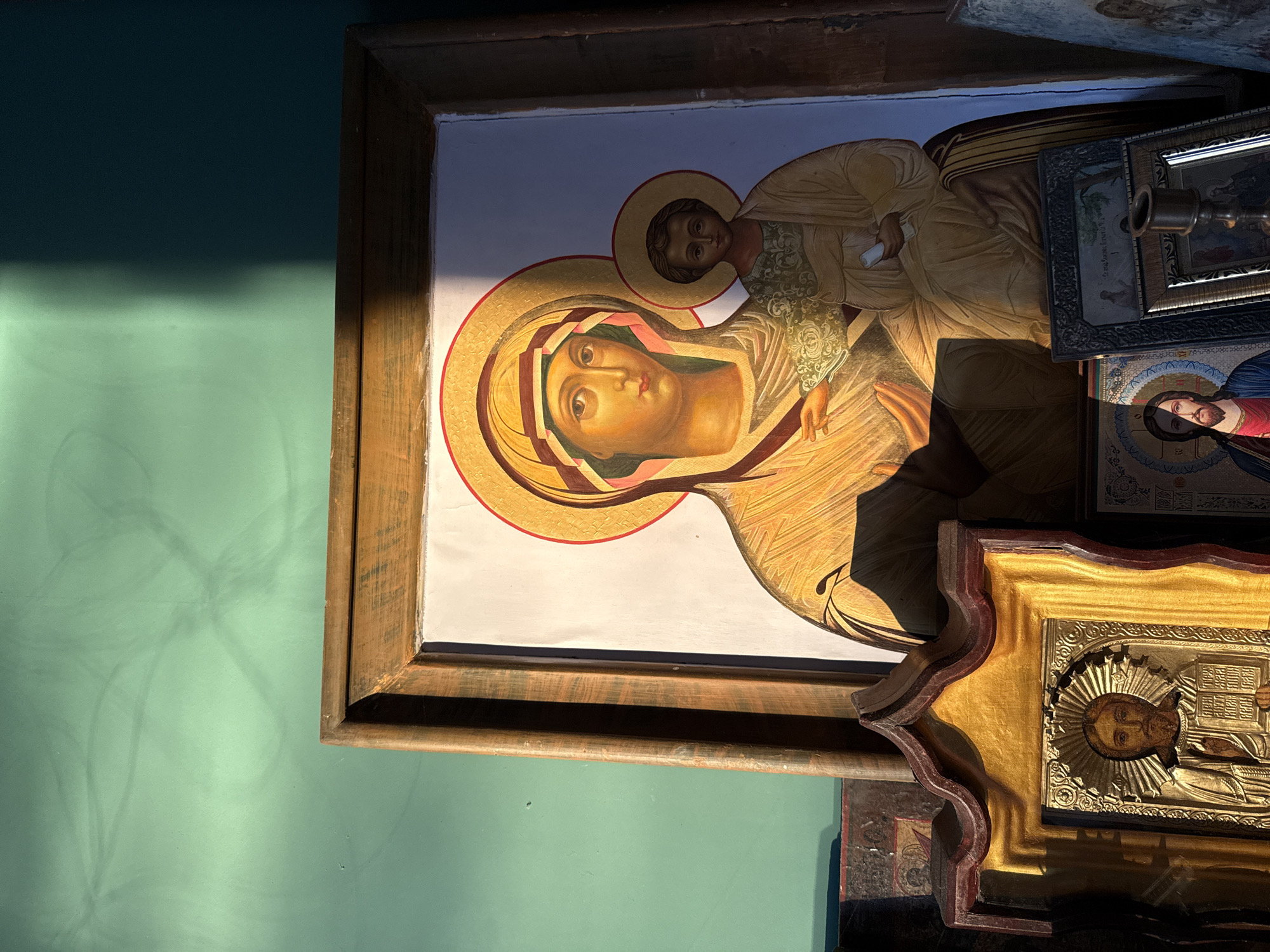 The image depicts a collection of religious icons displayed against a textured, light green wall. The primary icon, centrally positioned, is a large painting of the Virgin Mary holding the infant Jesus. Both figures have halos, and Mary is adorned in traditional religious attire with golden and yellow hues. The icon is housed in a rough, wooden frame.
To the left and in the foreground, a smaller, ornately carved golden icon is visible, featuring a depiction of Jesus Christ. Other icons and religious artifacts are partially visible in the background and to the right, suggesting a devotional or ecclesiastical setting. Sunlight casts strong shadows across the scene, highlighting the texture of the wall and creating a dramatic interplay of light and dark on the icons. The overall impression is one of quiet reverence and artistic tradition.
The scene is located in Votkinsk, Russia.