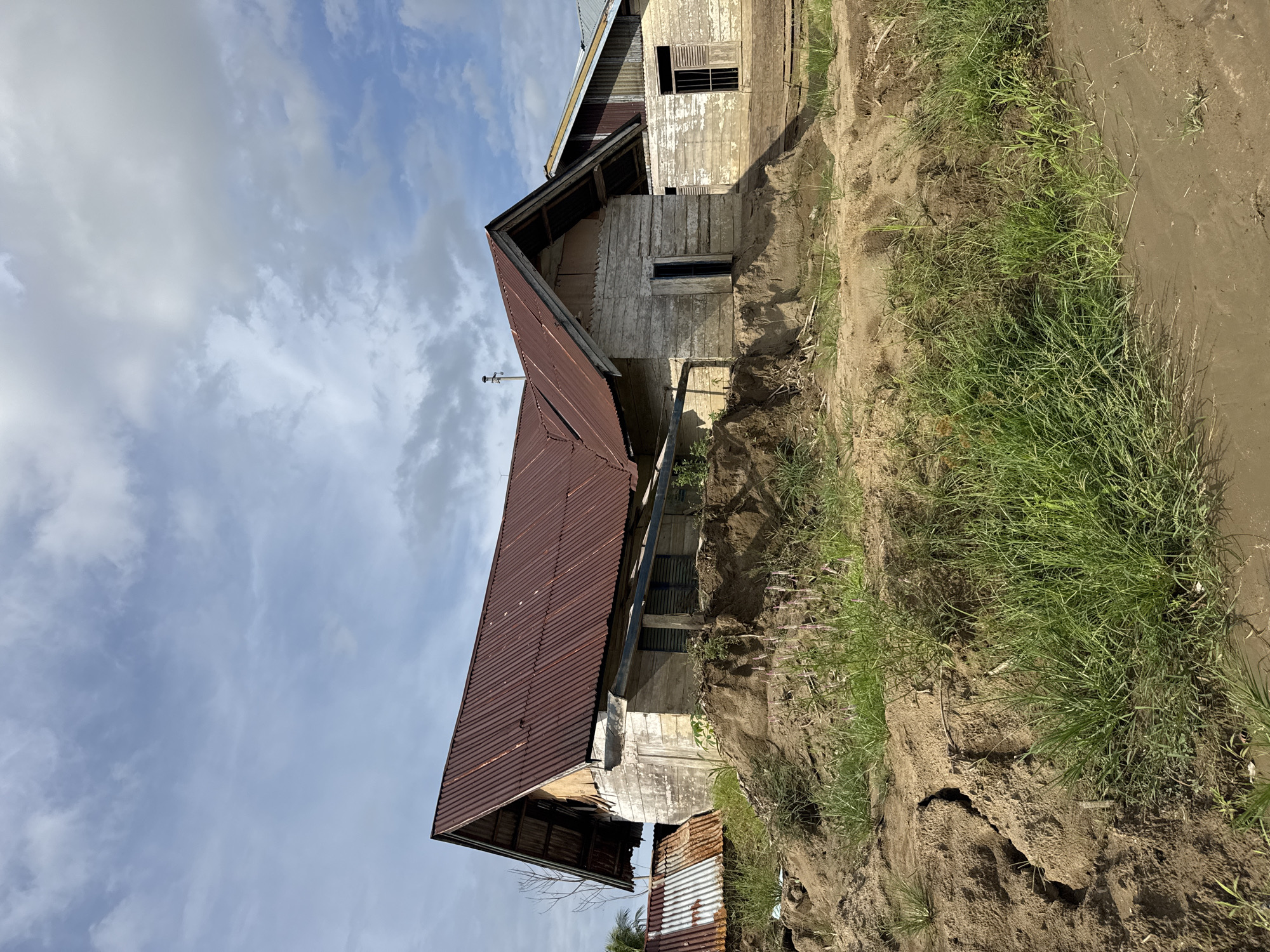 The image depicts a scene in Gampong Blang, Indonesia, showing two weathered wooden houses with corrugated metal roofs. The house in the foreground is partially built on a mound of earth with visible cracks, suggesting potential erosion or structural instability. Lush green grass grows around the base of the mound and in the foreground. The sky above is a mix of blue with scattered white clouds. The second house, visible to the right of the foreground structure, appears to be in similar condition with whitewashed wooden walls and a metal roof. The overall impression is one of rural simplicity and potential environmental challenges.
