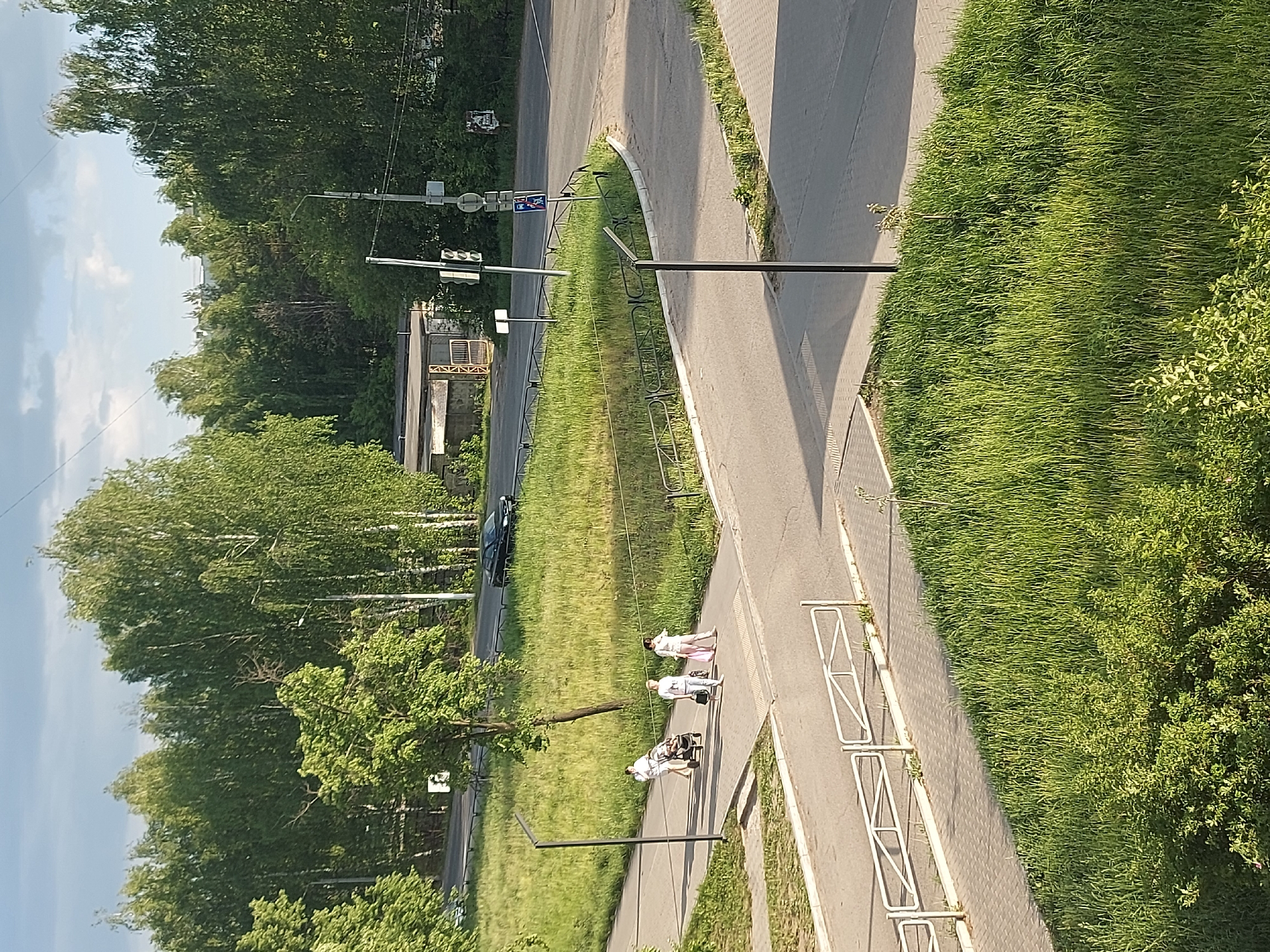 The image shows a street scene in Chaikovsky, Russia, viewed from above. A sidewalk runs along the right side of the frame, bordered by grass and trees. Three pedestrians are walking on the sidewalk: one pushing a stroller, followed by two women. A street runs parallel to the sidewalk, with trees and a building on the other side. There are also streetlights and traffic signs visible. A dark car is parked on the street. The overall environment appears to be a residential or suburban area during daylight.