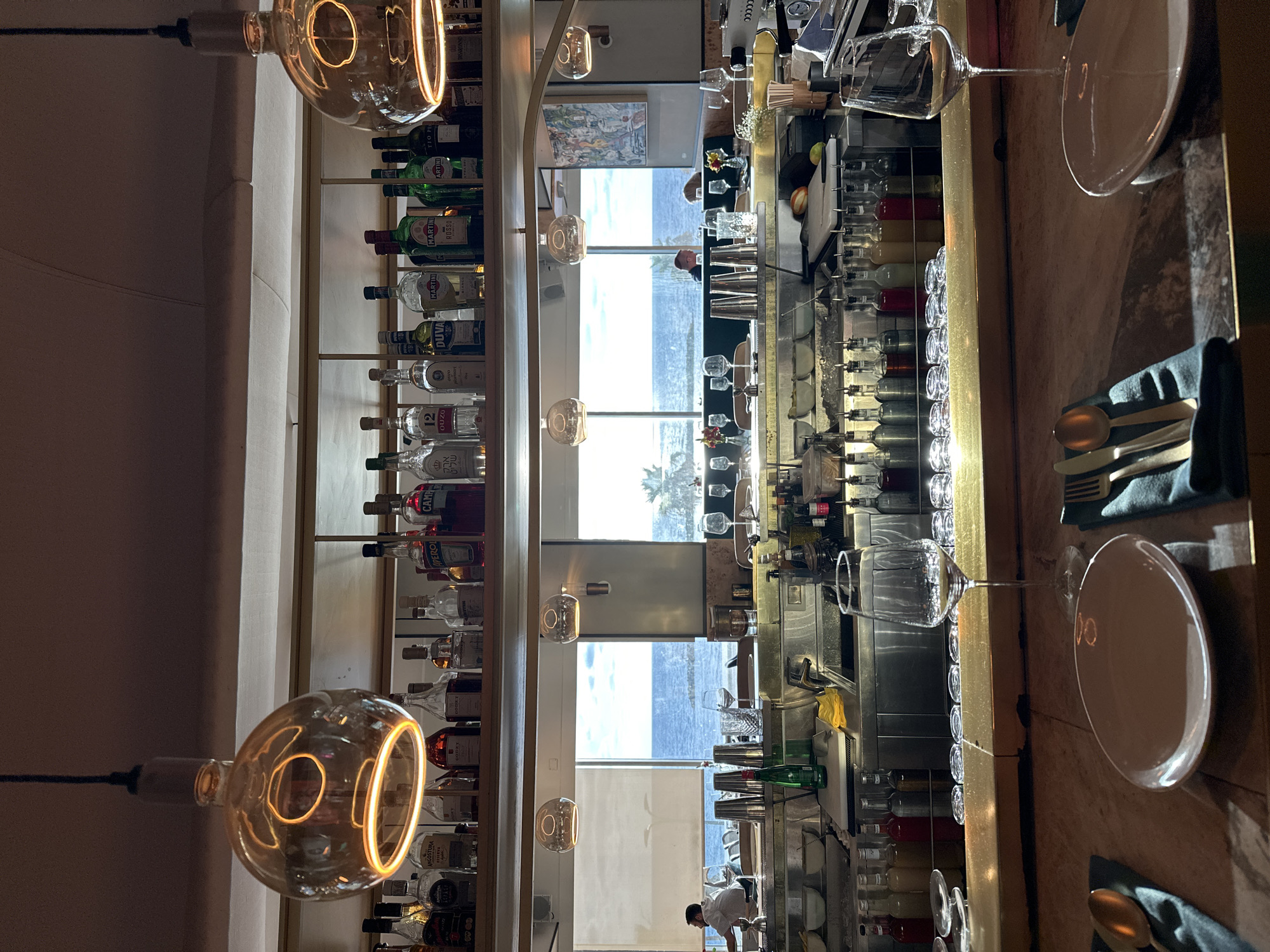 This image captures the interior of a bar or restaurant, likely in Tel-Aviv, Israel, judging by the coastal view visible through the large windows. The primary focus is on the bar setup, which features shelves stocked with a variety of liquor bottles, including brands like Martini, Aperol, Campari, and Angostura. A row of small liquor bottles is arranged along the top of the bar.
The bar counter is visible, with set tables featuring plates, cutlery, and napkins. Wine glasses are strategically placed, indicating a setting for dining. The backdrop reveals a scenic ocean view with a visible coastline and sky.
Hanging light fixtures with large, decorative bulbs are present, casting a warm glow. Towards the back, a few people can be seen in the reflection from the bar mirror.