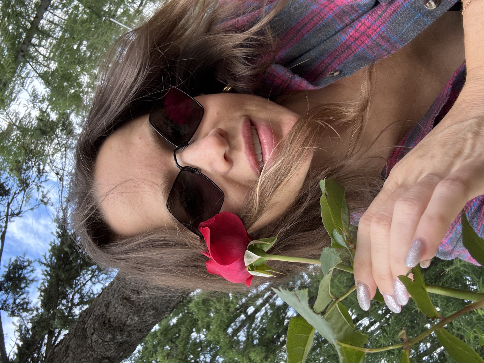 The image is a close-up, selfie-style photograph of a woman holding a red rose. She is wearing sunglasses and a plaid shirt. The background appears to be outdoors with trees and a cloudy sky. There is no indication of a specific location in Moscow, Russia.