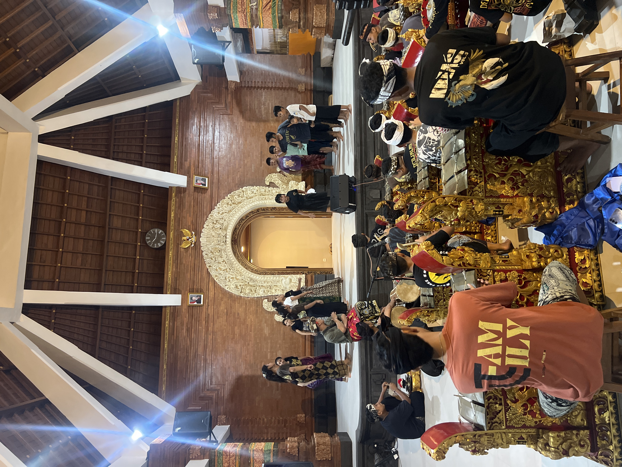 The image depicts a scene inside a building in Sukawati, Indonesia. On one side, a group of people are standing near a decorated archway. On the opposite side, individuals are seated, seemingly playing musical instruments, with ornate, golden stands. The interior features wooden walls and a high ceiling with white support beams. A clock is visible on the wall. There is also a speaker to the lower left of the image.
