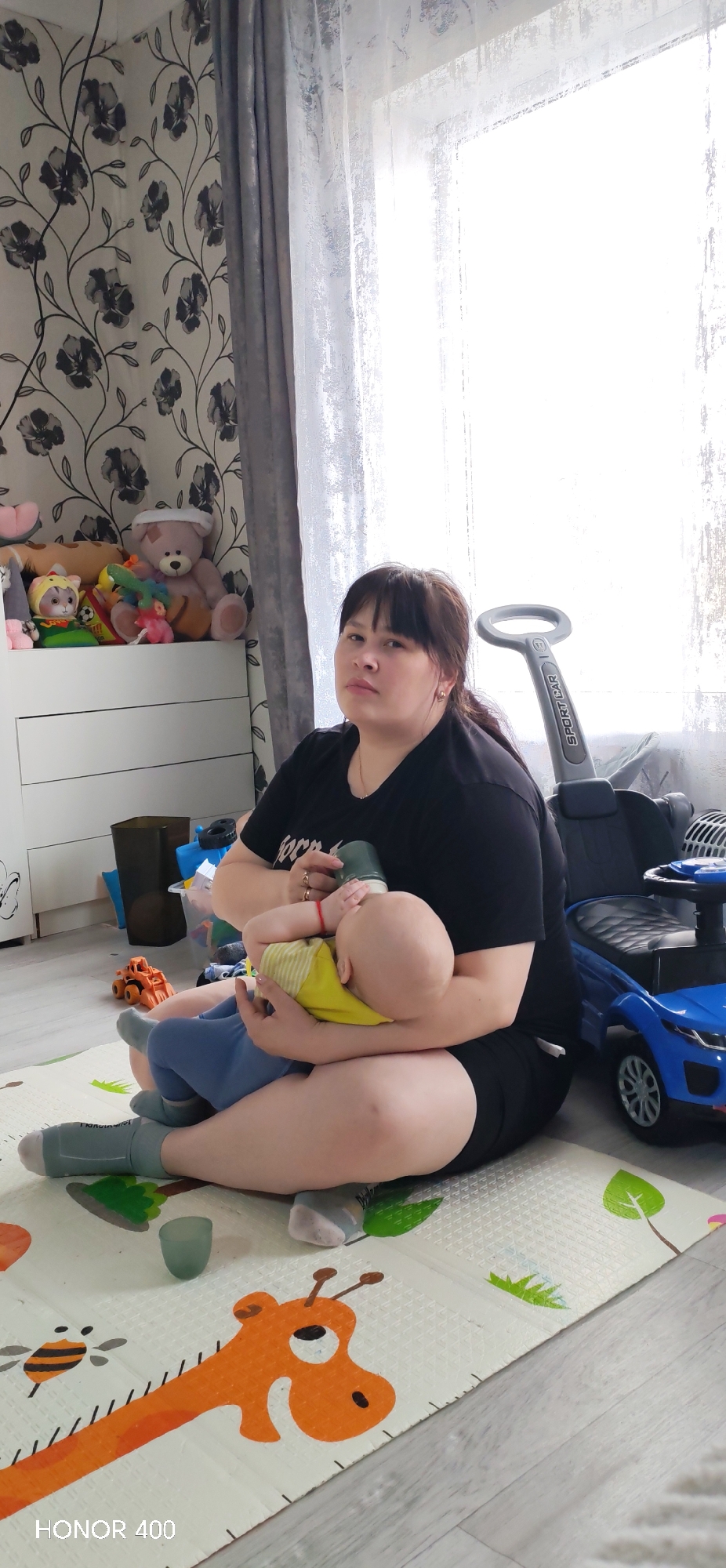 This full-length vertical shot depicts a woman bottle-feeding a baby on the floor of a brightly lit room in Пушкари, Russia. The woman, with dark hair and bangs, wears a black t-shirt and black shorts, with grey socks. She is seated cross-legged on a colorful playmat, holding the baby who is dressed in a yellow top and blue bottoms. Her expression is neutral as she looks towards the camera while holding a green baby bottle to the infant's mouth.
The setting appears to be a child's room or play area. To the left, the wall is adorned with wallpaper featuring black floral patterns on a light background. A white dresser or cabinet is visible behind the woman, piled with various plush toys, including a large brown teddy bear with a bandage on its head. To the right, a large window allows bright daylight into the room, covered by dark grey curtains and sheer white curtains. A blue toy car or ride-on toy, featuring a gray handle with "SPORT CAR" text, is positioned near the window. The playmat beneath the subjects has cheerful illustrations, including an orange giraffe and a bee. A small, dark green cup rests on the playmat near the woman's feet. The floor surrounding the playmat appears to be light-colored wood or laminate.
The time of day is daytime, evidenced by the bright light from the window. The woman's emotion is calm and attentive. The text "HONOR 400" is visible in the bottom left corner of the image.